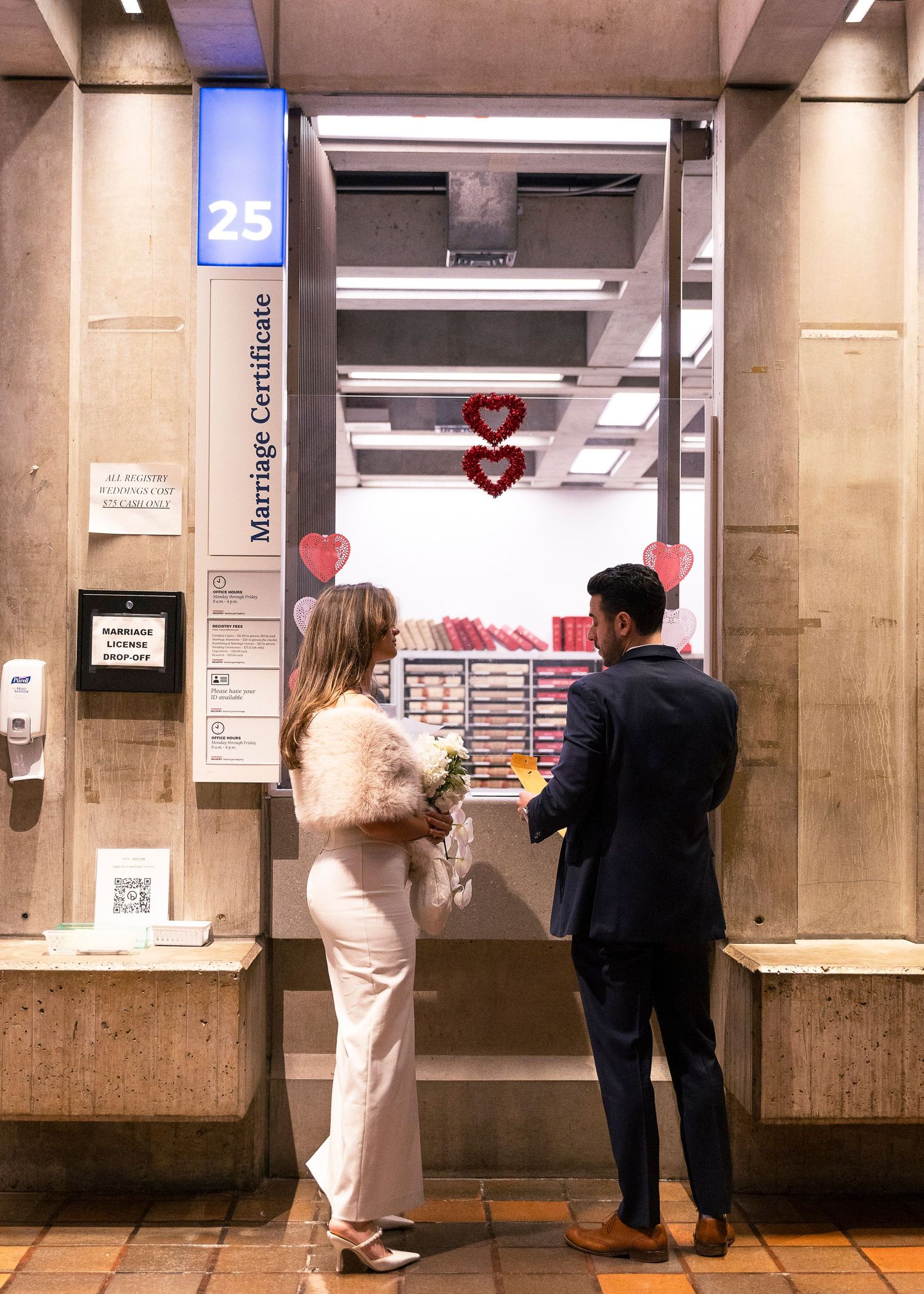 Couple in wedding attire at a Marriage Certificate counter 25, woman holding bouquet, decorated with red hearts.