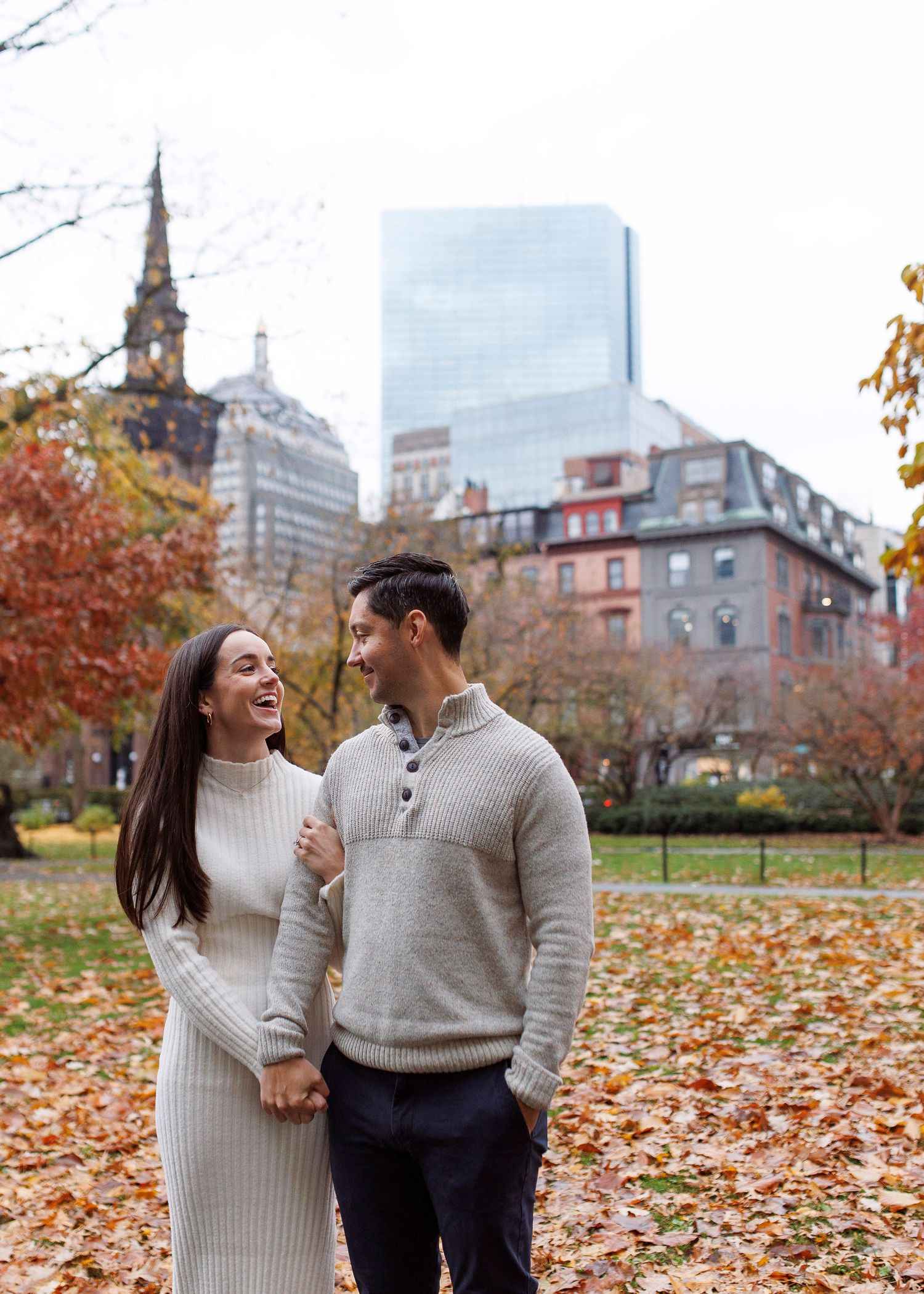 A couple in matching beige sweaters share a joyful moment in a city park during autumn.
