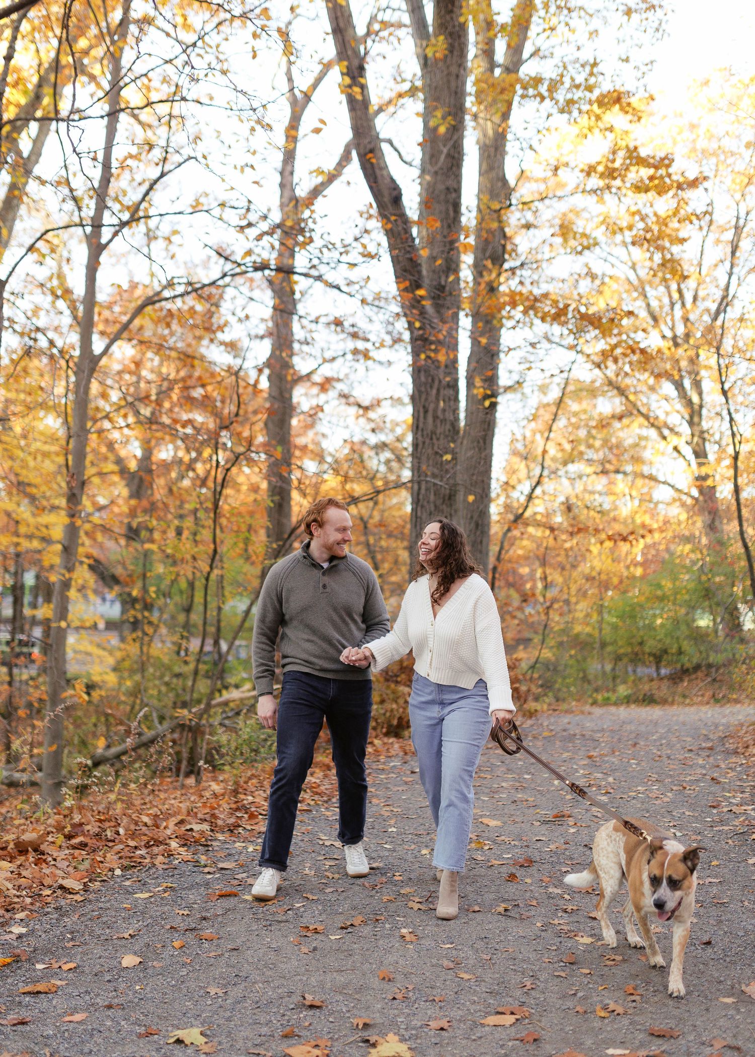 A couple walks their dog along a tree-lined path covered in fallen autumn leaves on a sunny afternoon.