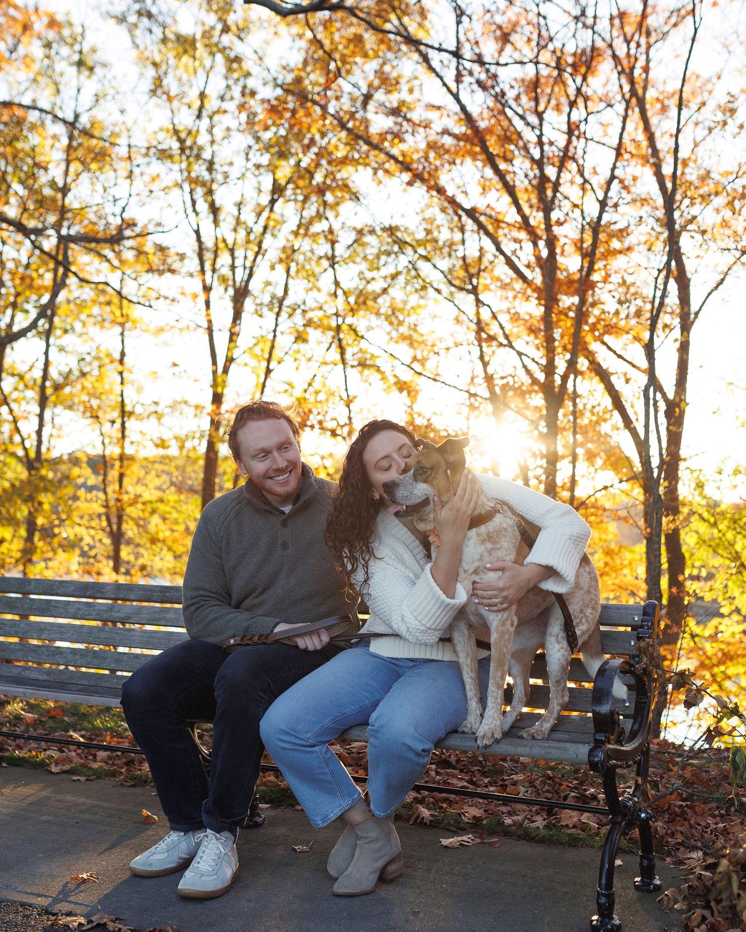 Couple sitting on a park bench with their golden dog during autumn sunset, surrounded by colorful fall foliage.