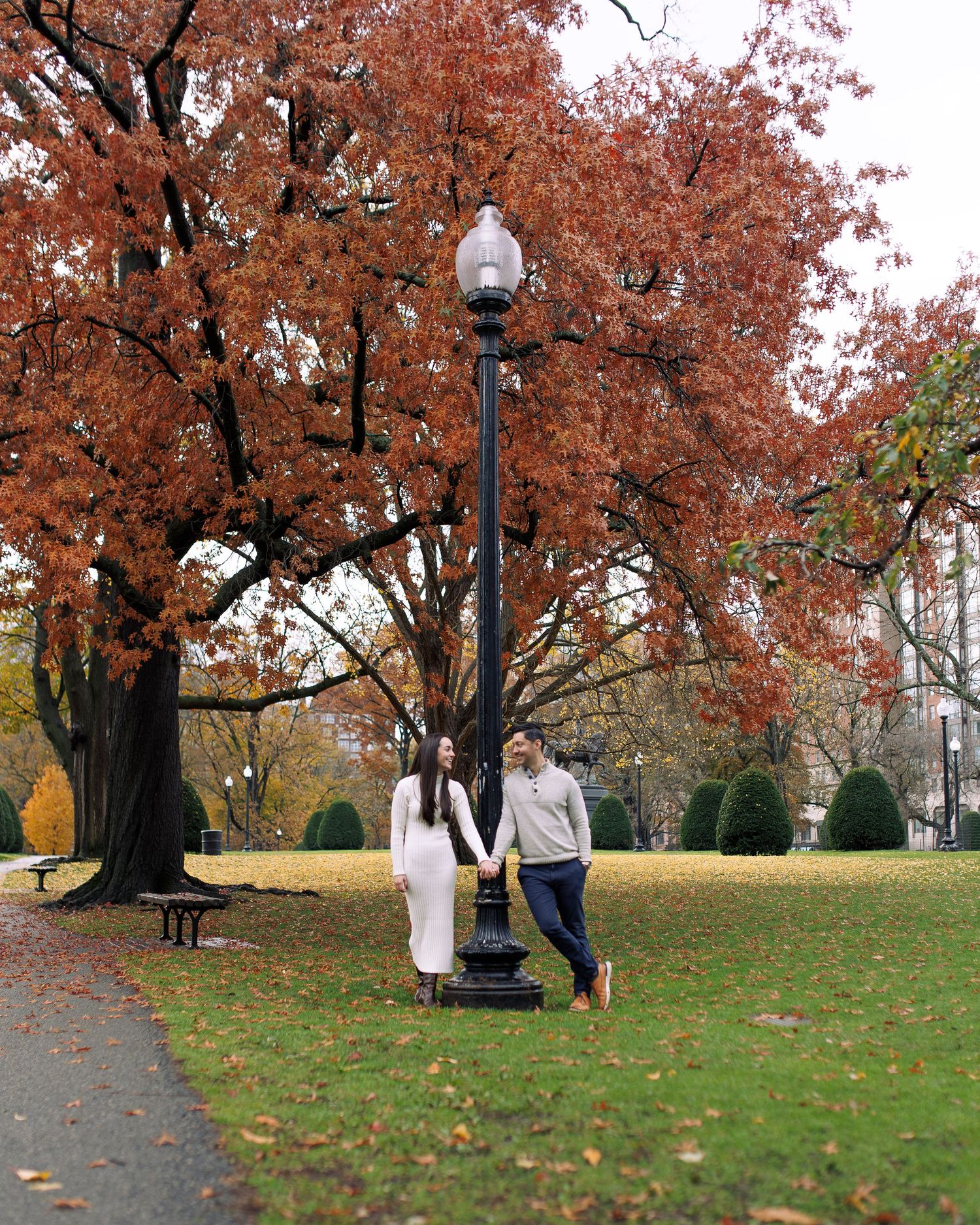 Couple dancing and twirling under vibrant orange autumn trees in a park setting with fallen leaves.