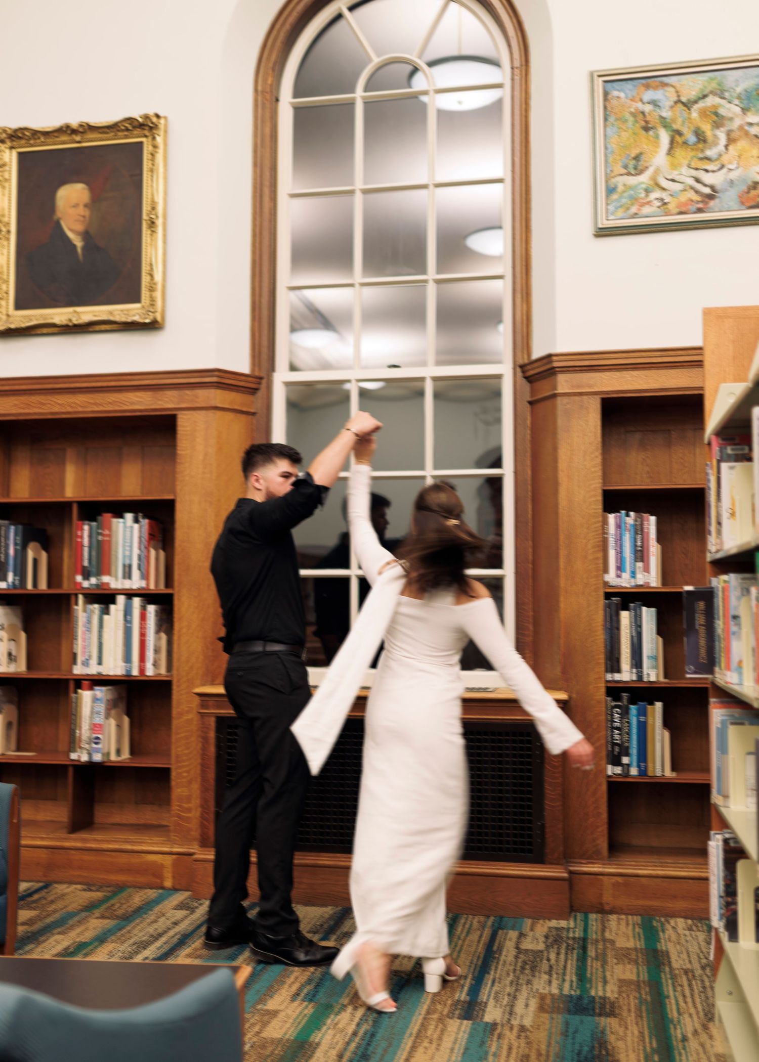 A couple dances together in an elegant library room with wooden bookshelves and a tall arched window.