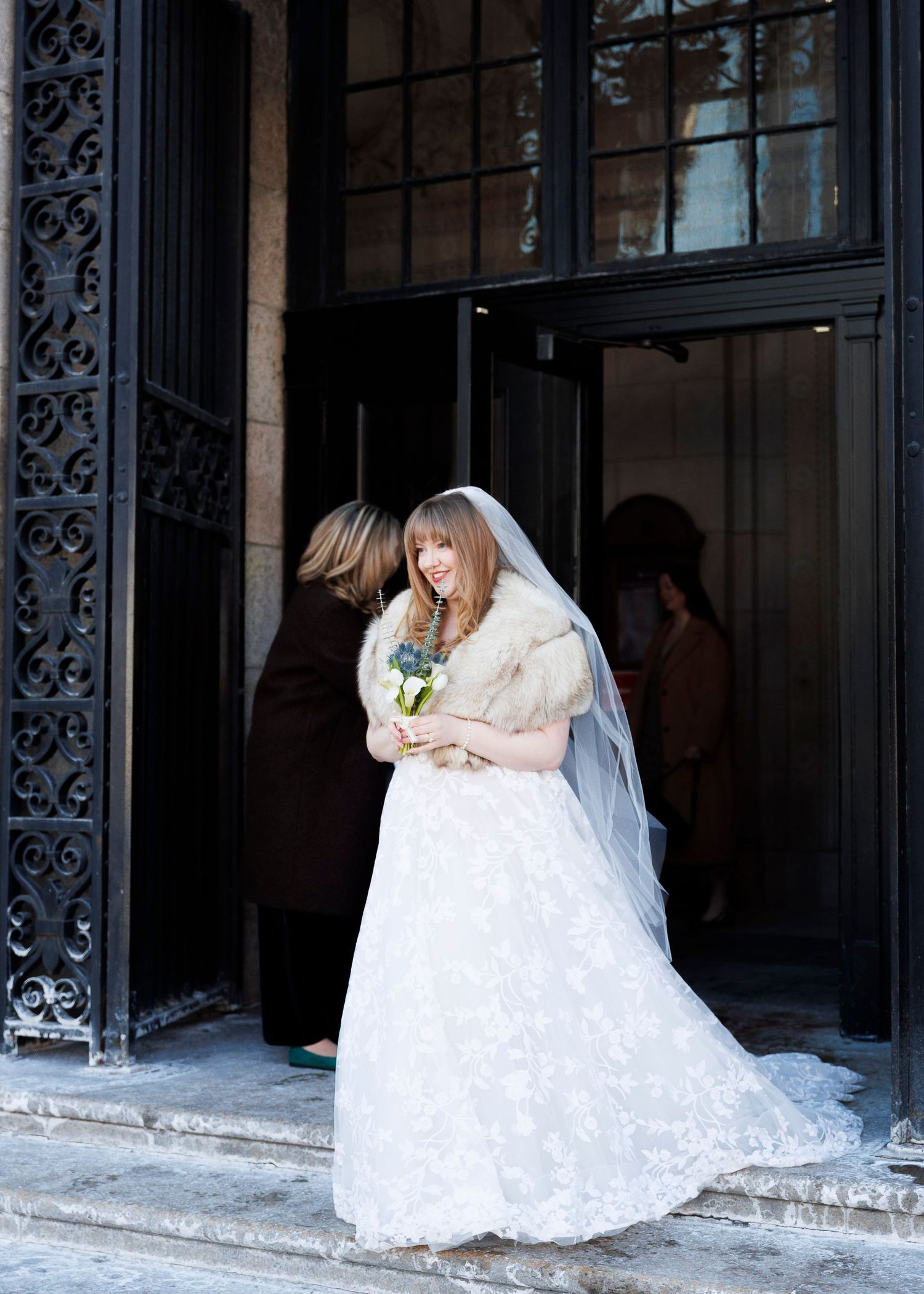 A bride in a white lace wedding dress sits on ornate stone steps in front of a decorative iron gate.