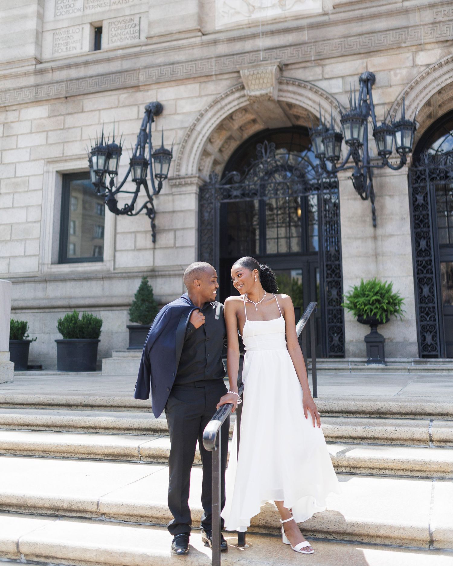 Couple in wedding attire poses on stone steps outside elegant building, groom in navy suit, bride in white dress.