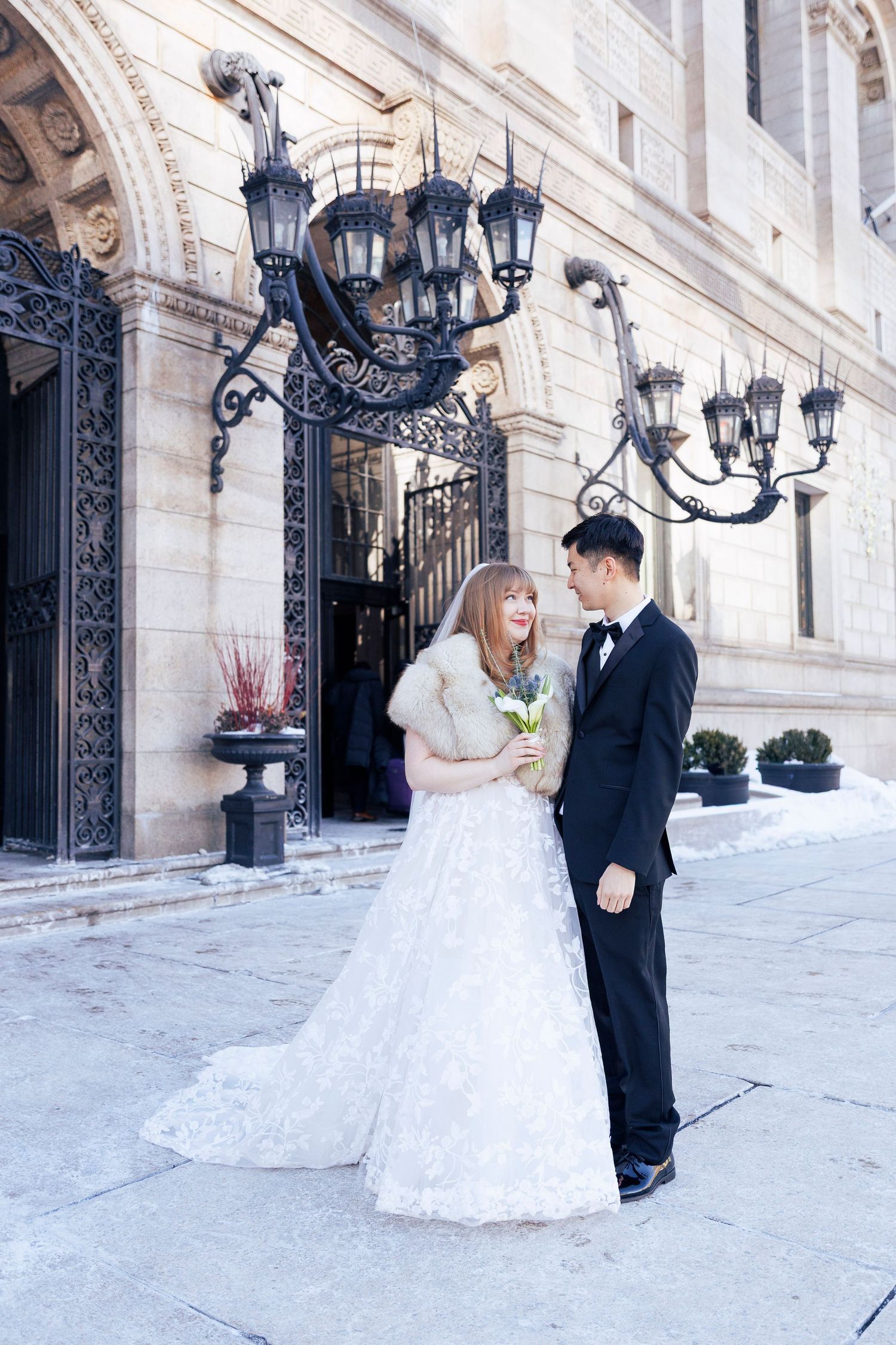 A bride and groom share a romantic moment outside an elegant building with ornate lamp posts and stone architecture.