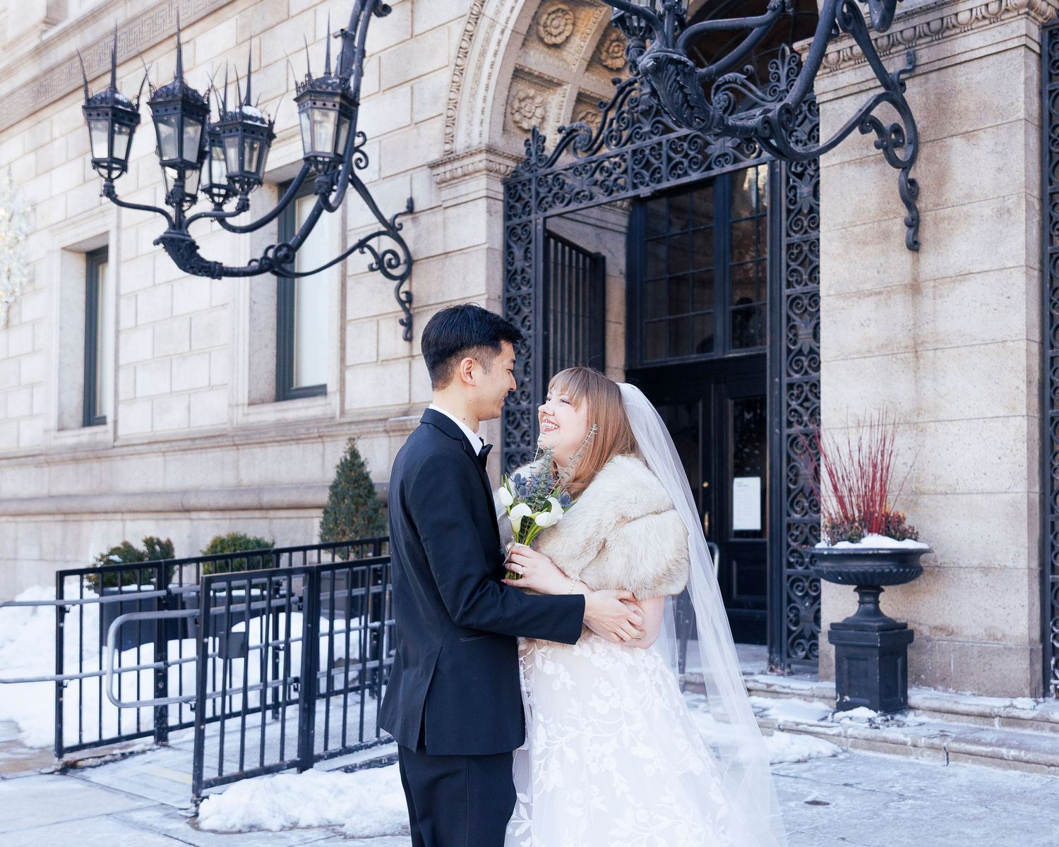 A couple in wedding attire poses together in a snowy winter setting with elegant architecture and ornate gates.