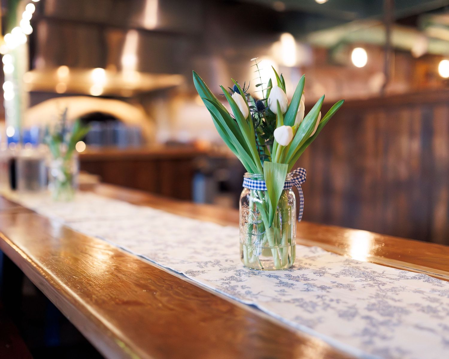 A rustic wooden bar counter decorated with a floral runner and white tulips in glass vases creates an elegant ambiance.