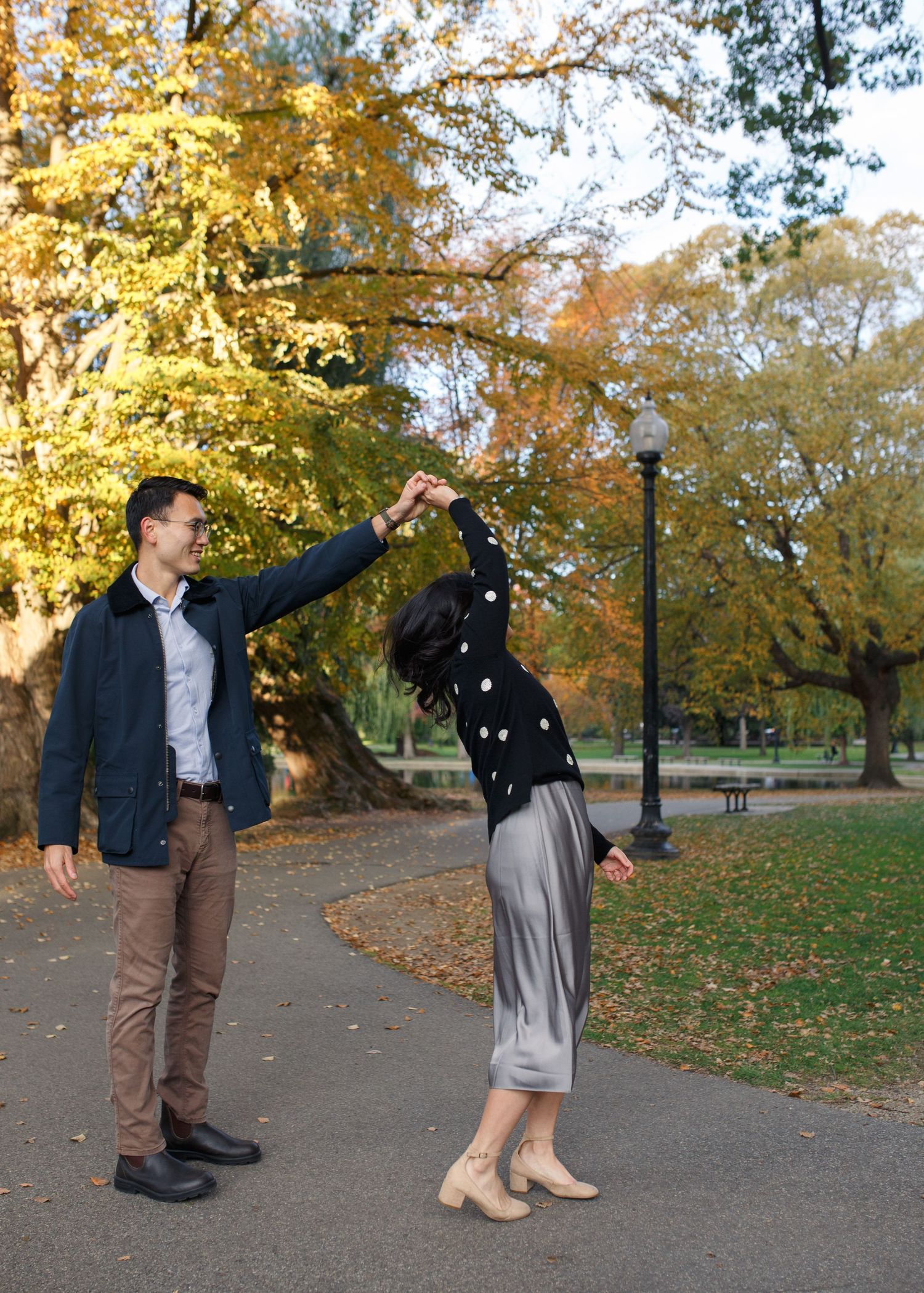 A couple dances playfully on a park path surrounded by golden fall foliage.