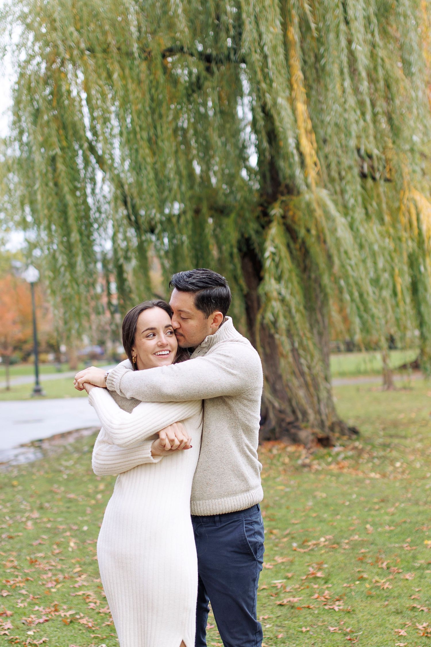 A couple in autumn attire embraces beneath weeping willow trees in a park setting.