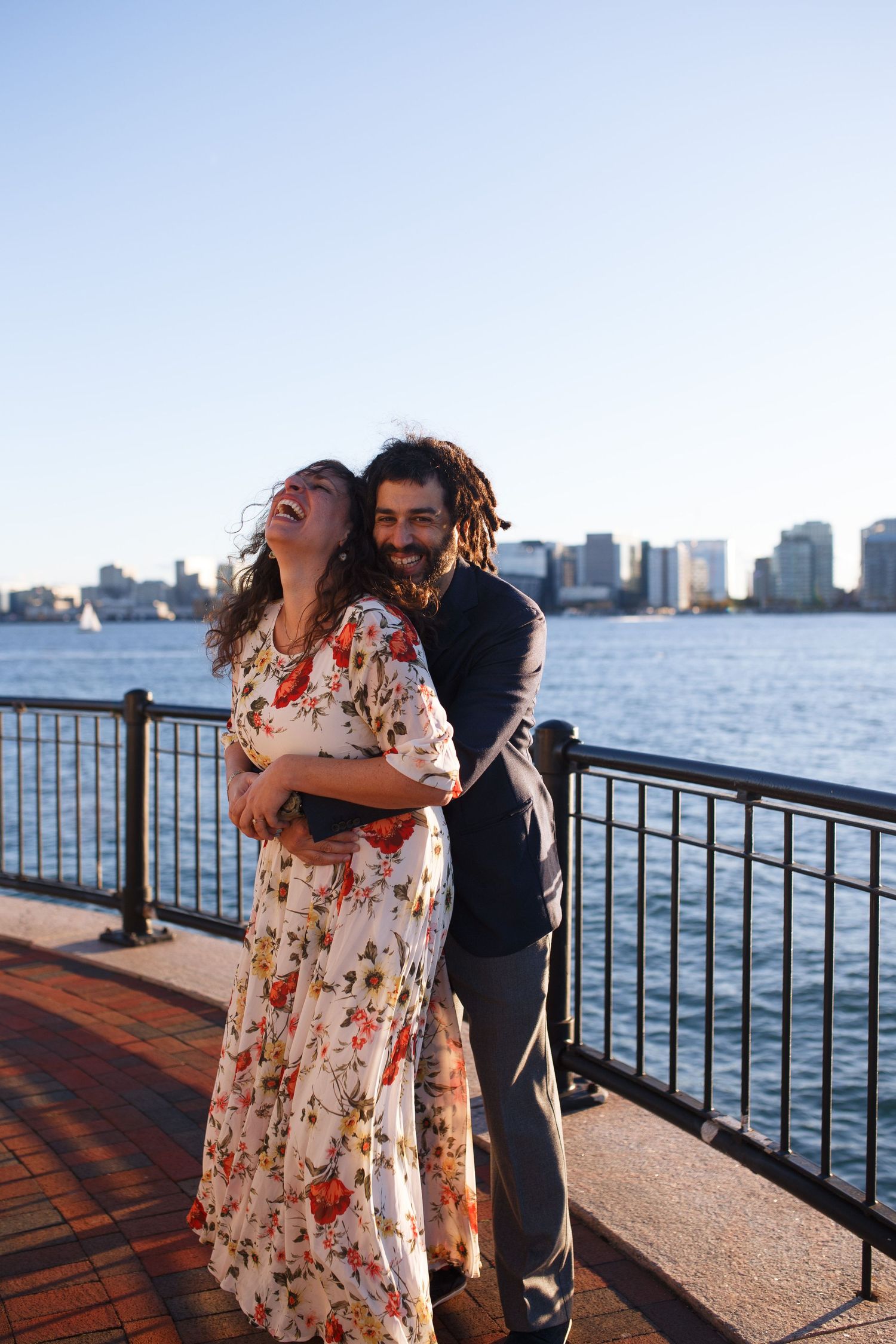 A romantic couple embraces on a waterfront boardwalk with city skyline in the background.