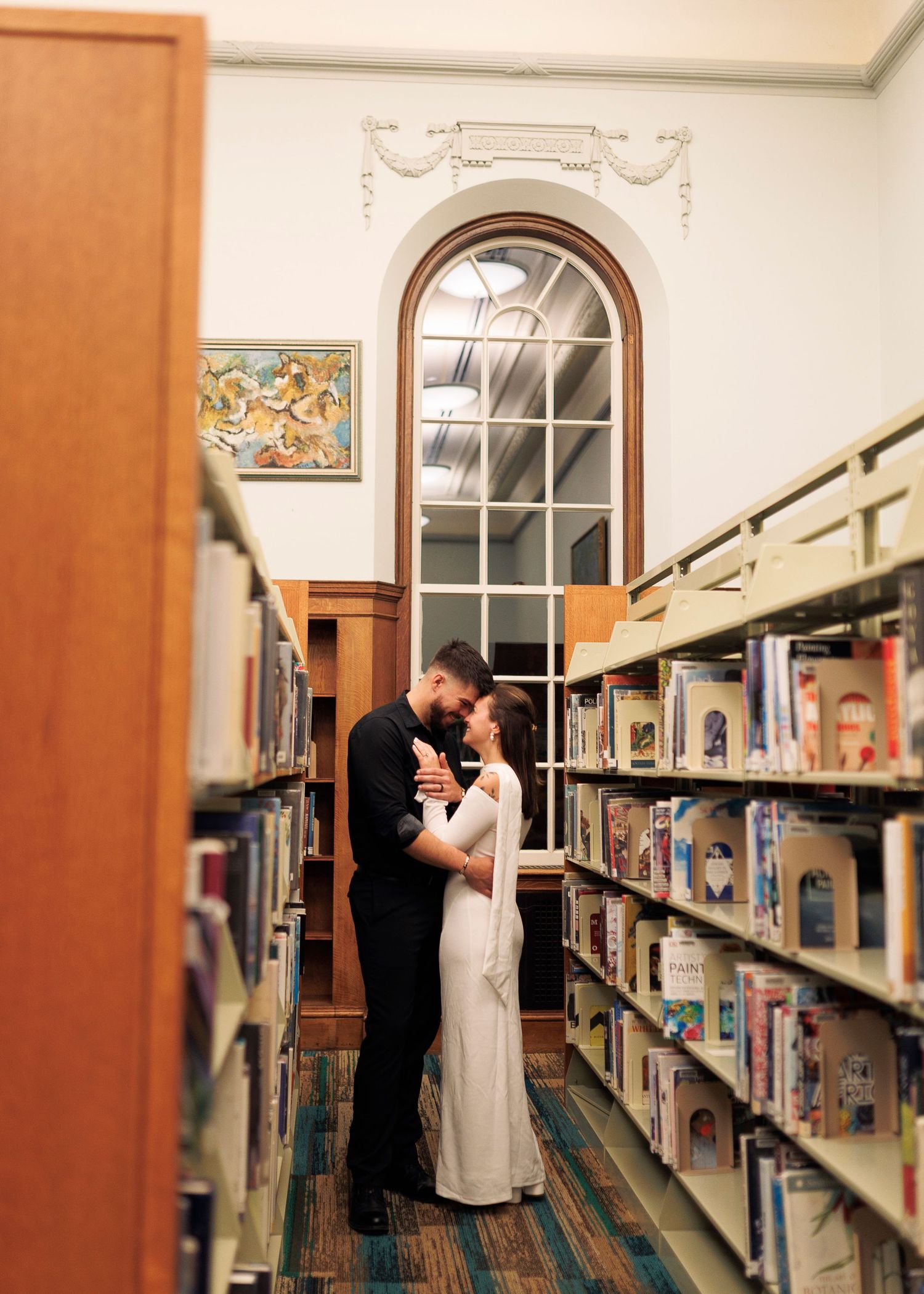 A couple shares a romantic moment between library bookshelves, creating an intimate scene in a quiet library setting.