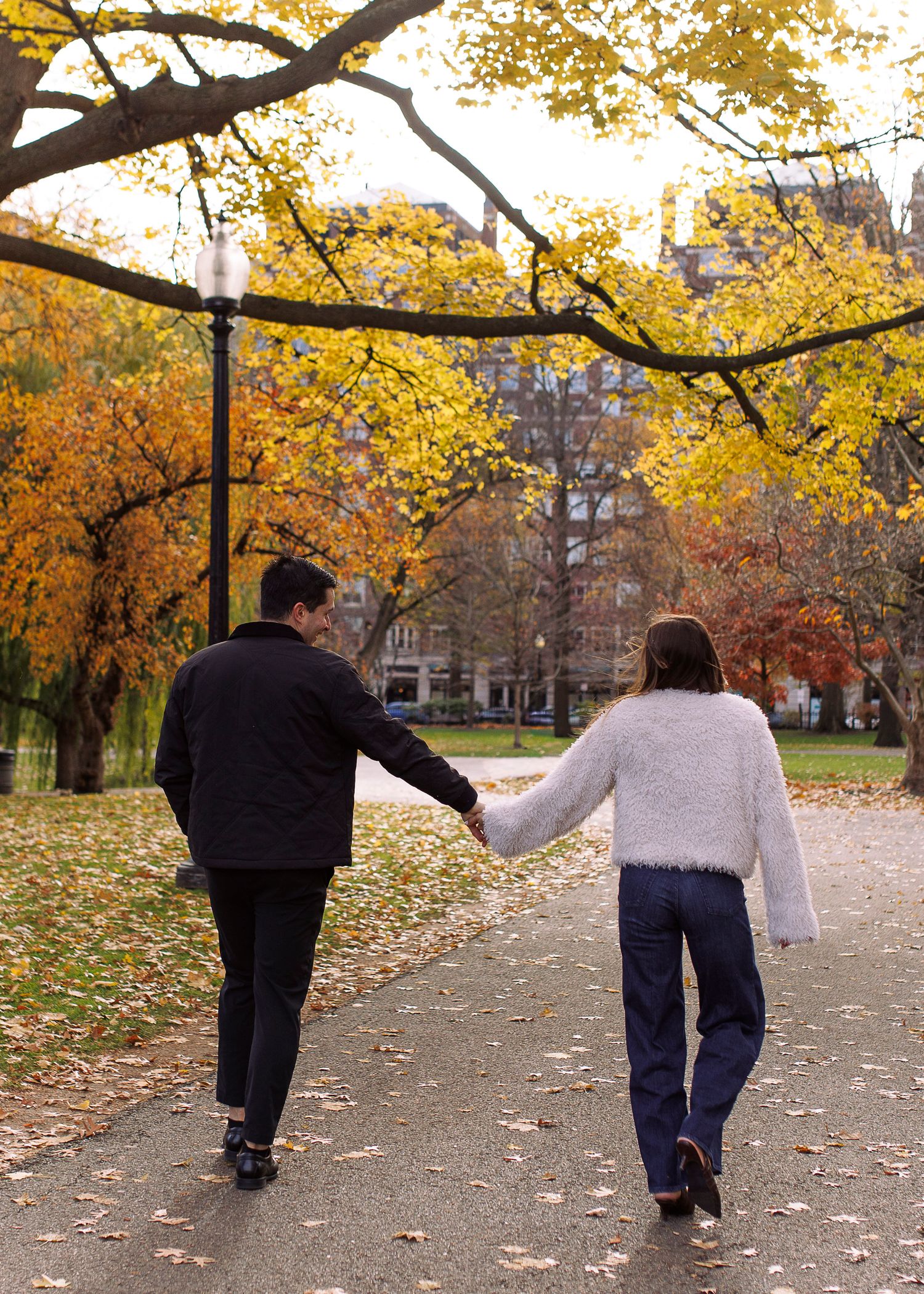 Two people walk along a tree-lined autumn path with fallen leaves while holding hands.