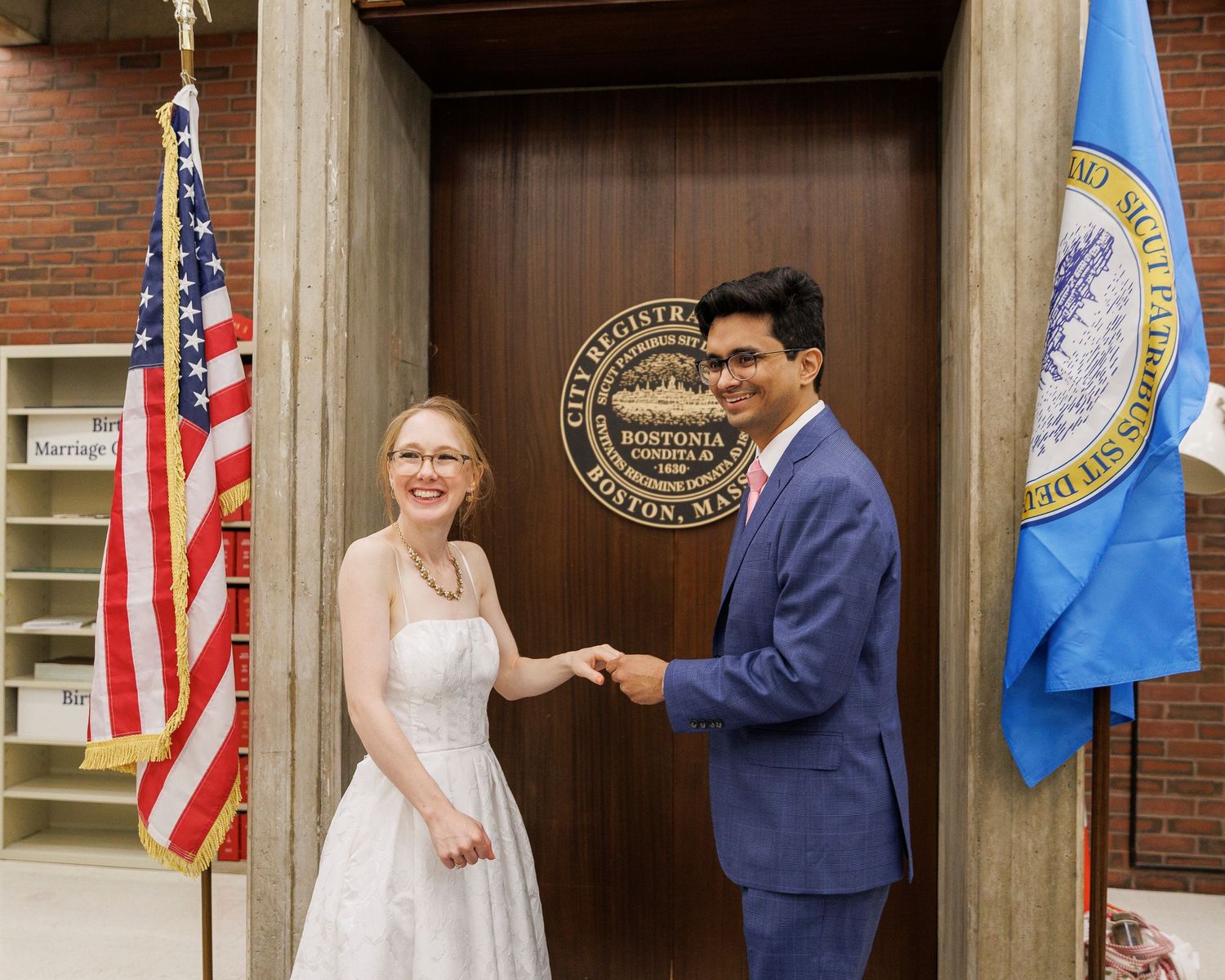 Couple holding hands at Boston Marriage Registry office, woman in white dress and man in blue suit, American and state flags.