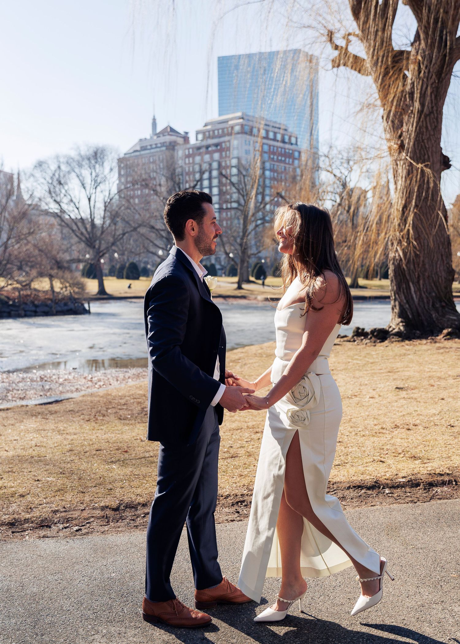 A couple in formal attire holds hands while standing on a path in Central Park with city buildings in the background.