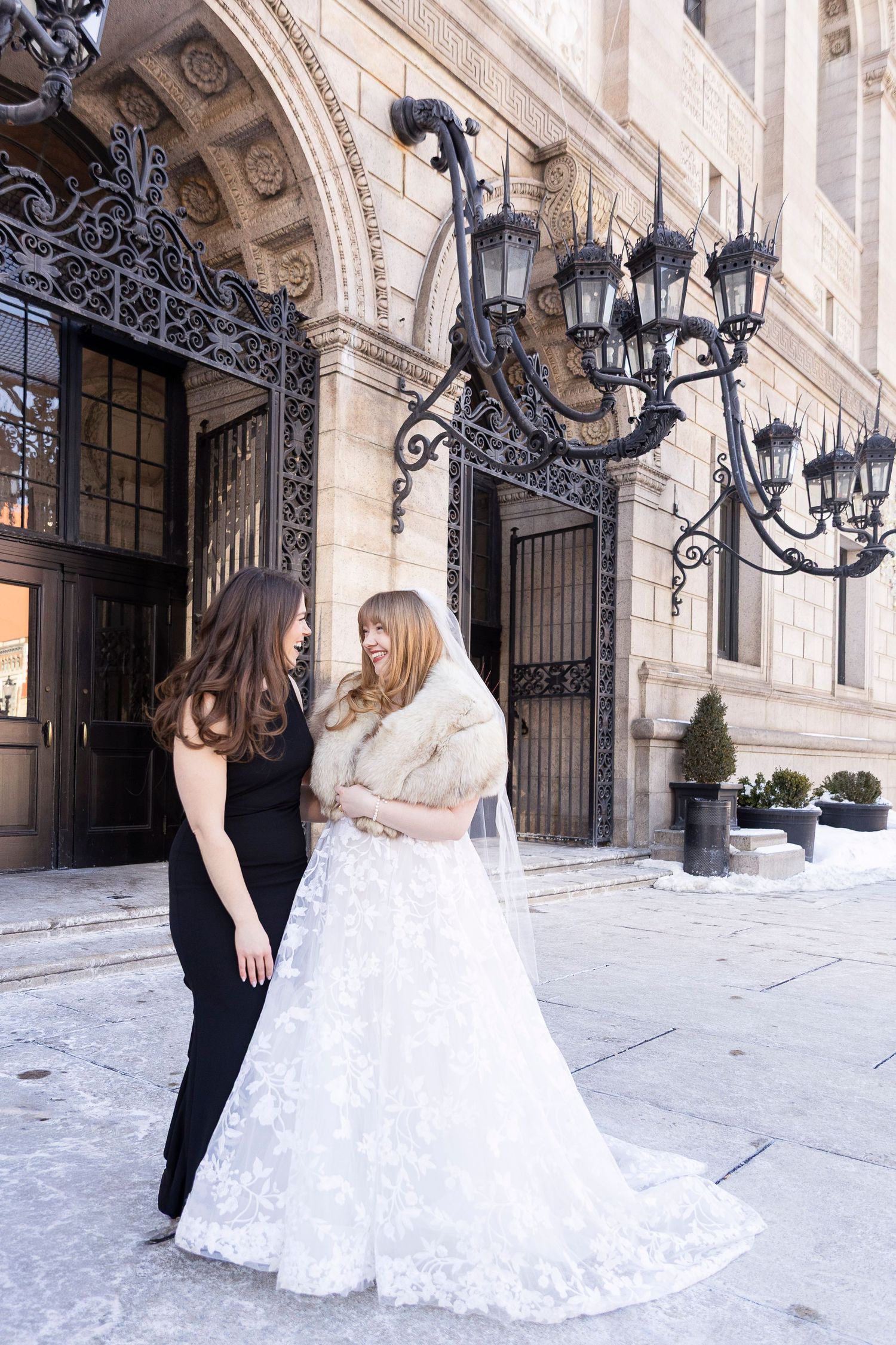 A couple in formal wedding attire stands outside an elegant building with ornate architecture and decorative lamp posts.