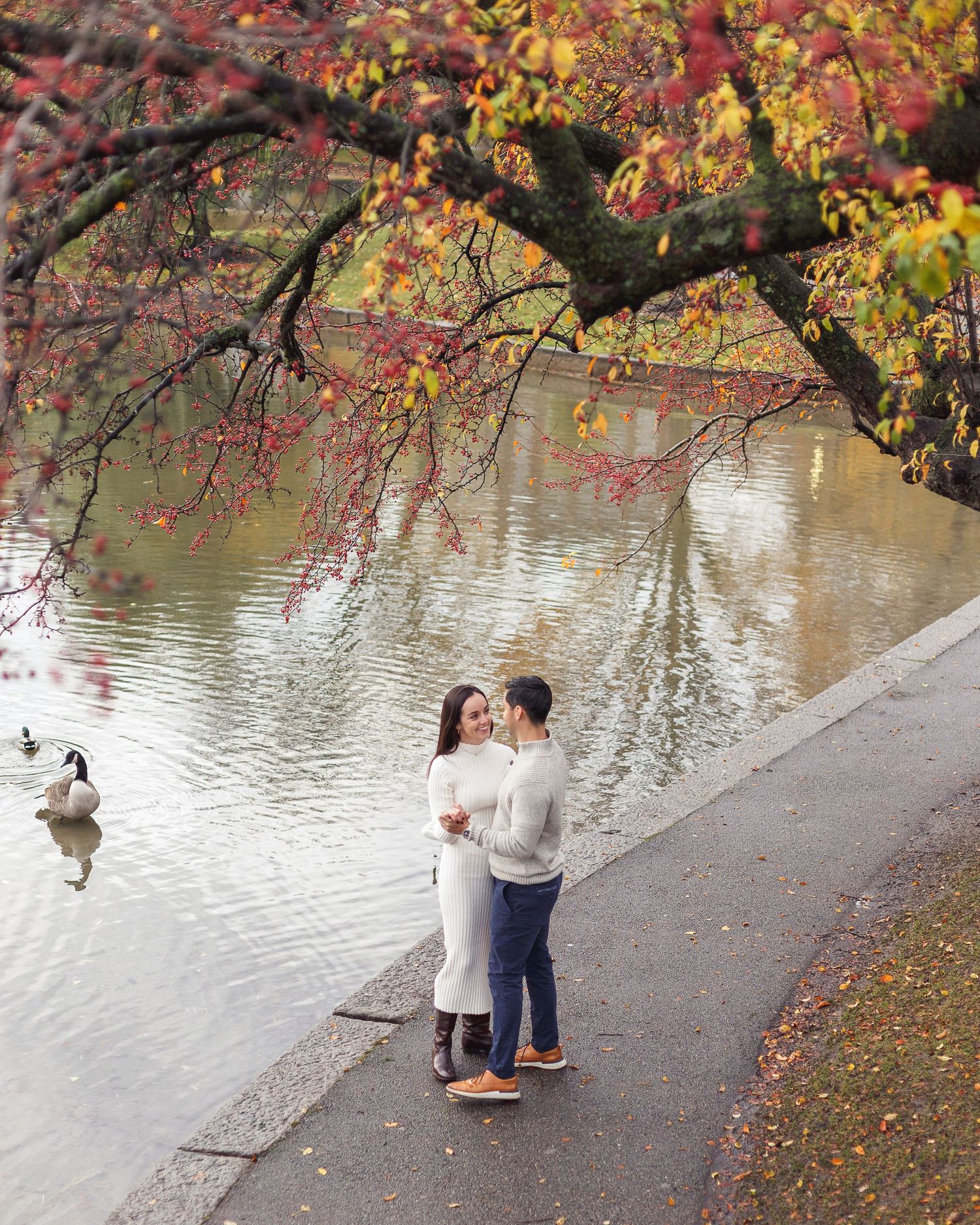 Two people stand by a lake in autumn with colorful fall foliage reflecting in the calm water.