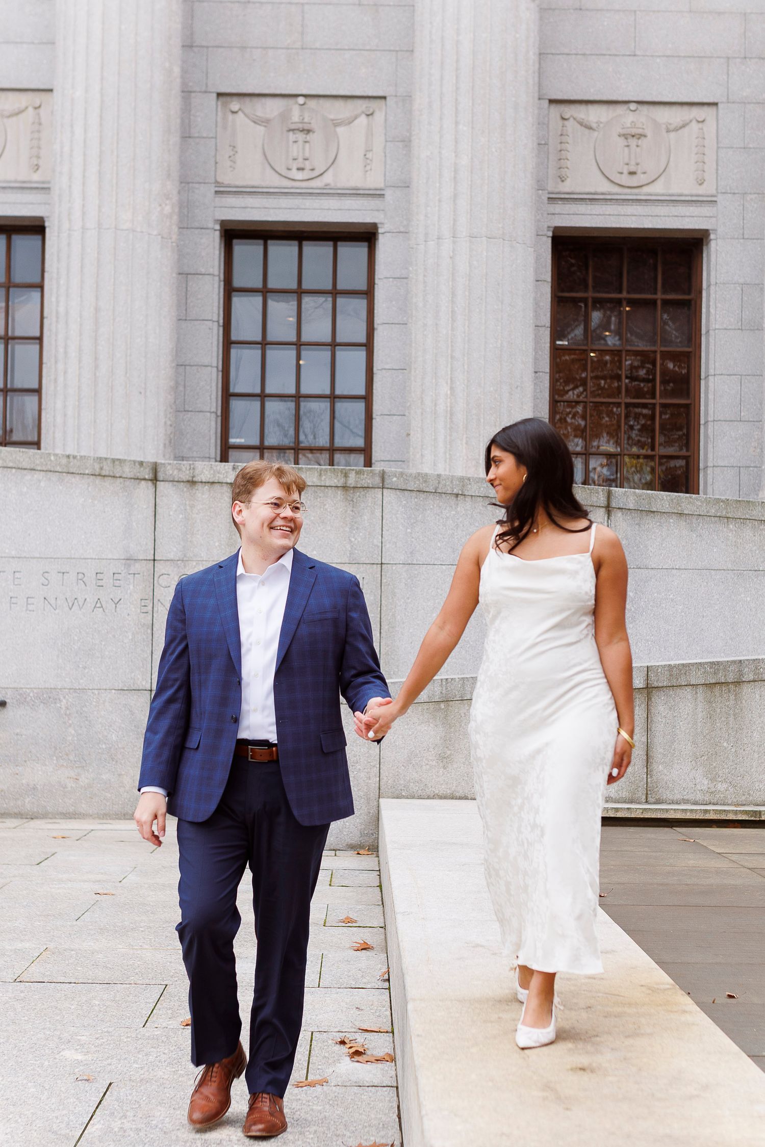 A couple in formal attire shares a playful moment on the steps of a grand stone building with columns.