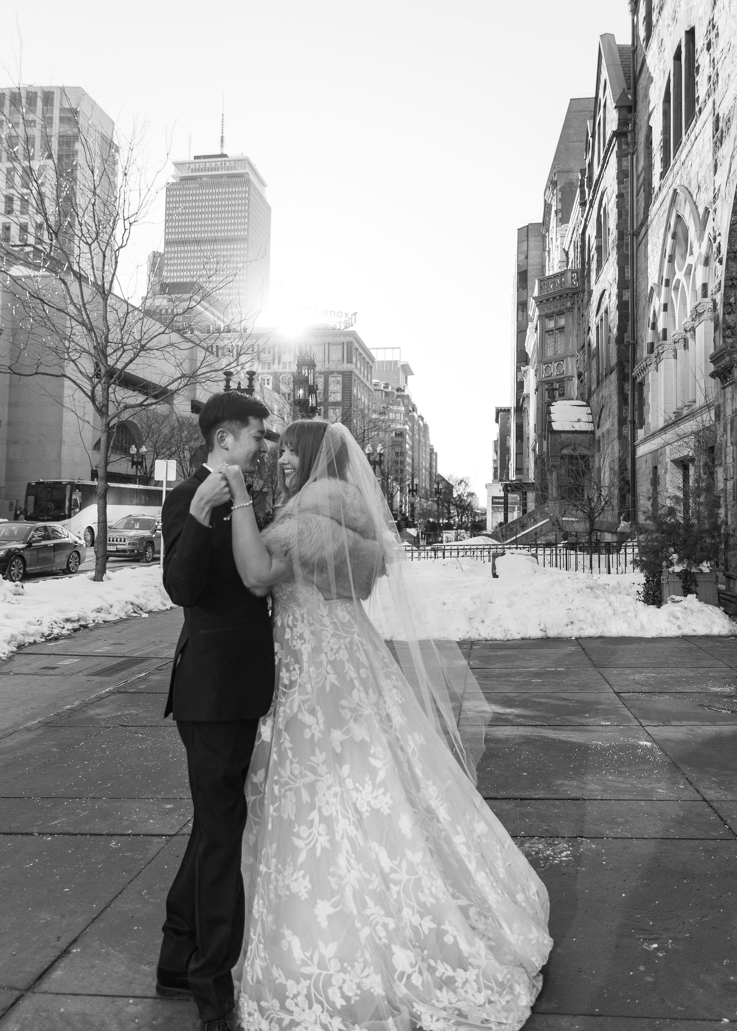 Black and white photo of a bride and groom embracing on a snowy city street, with sunlight glowing behind them.