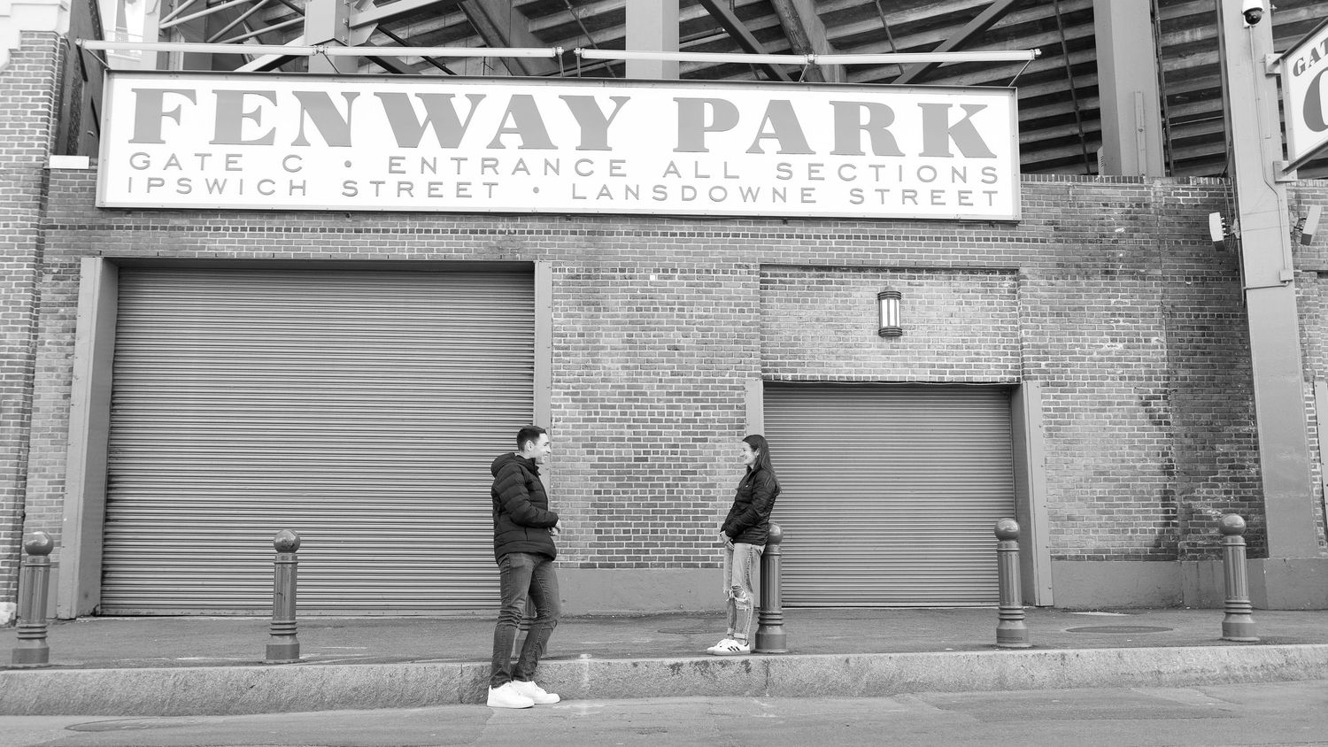 Two people stand outside Fenway Park gate C entrance on Ipswich and Lansdowne Street in black and white.