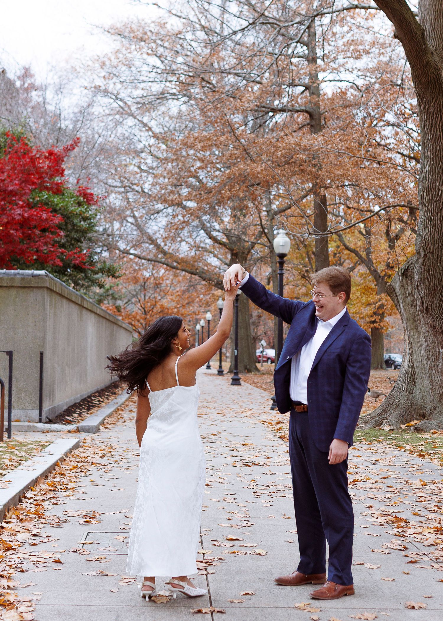 A couple playfully dances together on a walkway covered in autumn leaves during a fall photoshoot.