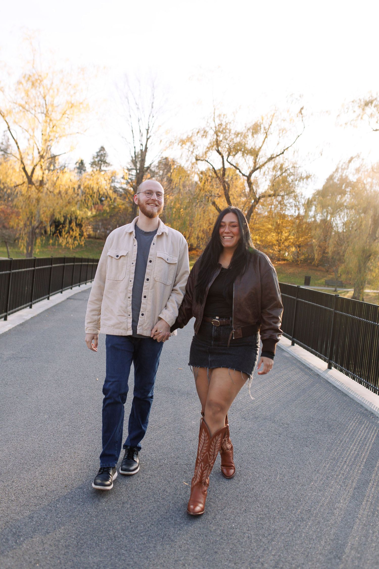 A couple walks together on a bridge path during autumn with fall foliage in the background.