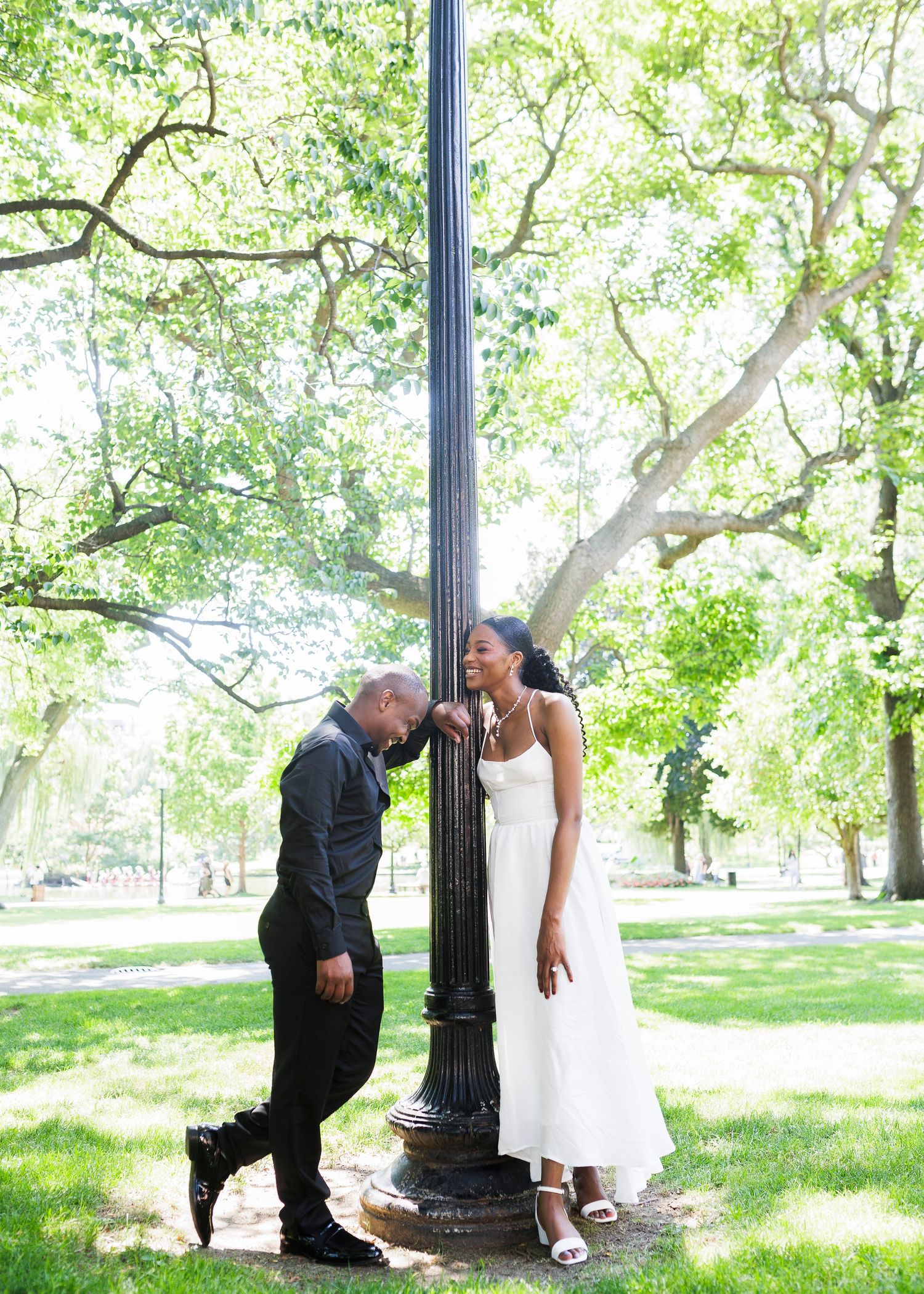 Couple in formal attire poses by a lamppost in a lush green park, woman in white dress smiles down at man.