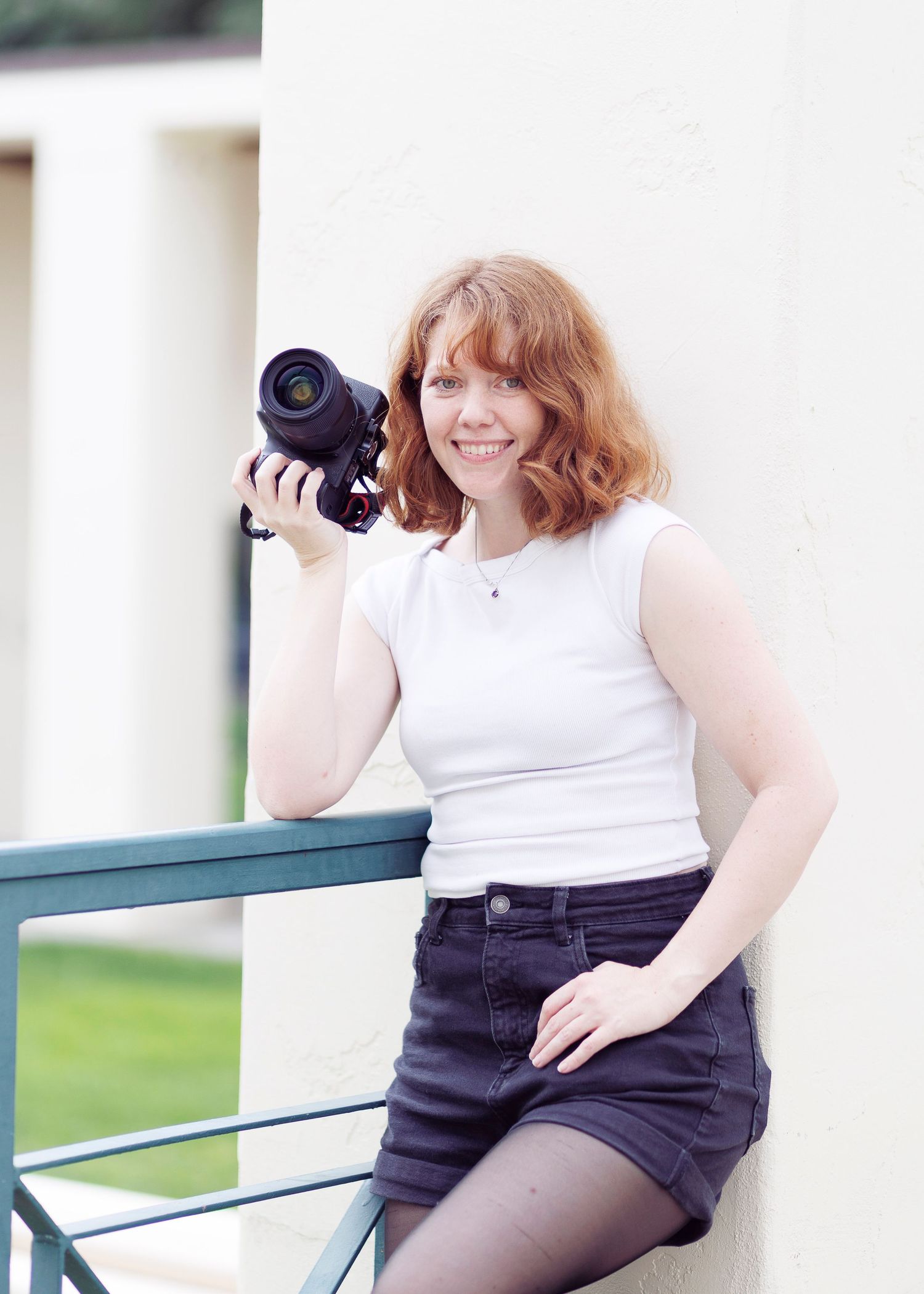 Smiling redhead woman holding a DSLR camera, wearing white tank top and denim skirt, leaning against a wall outdoors.