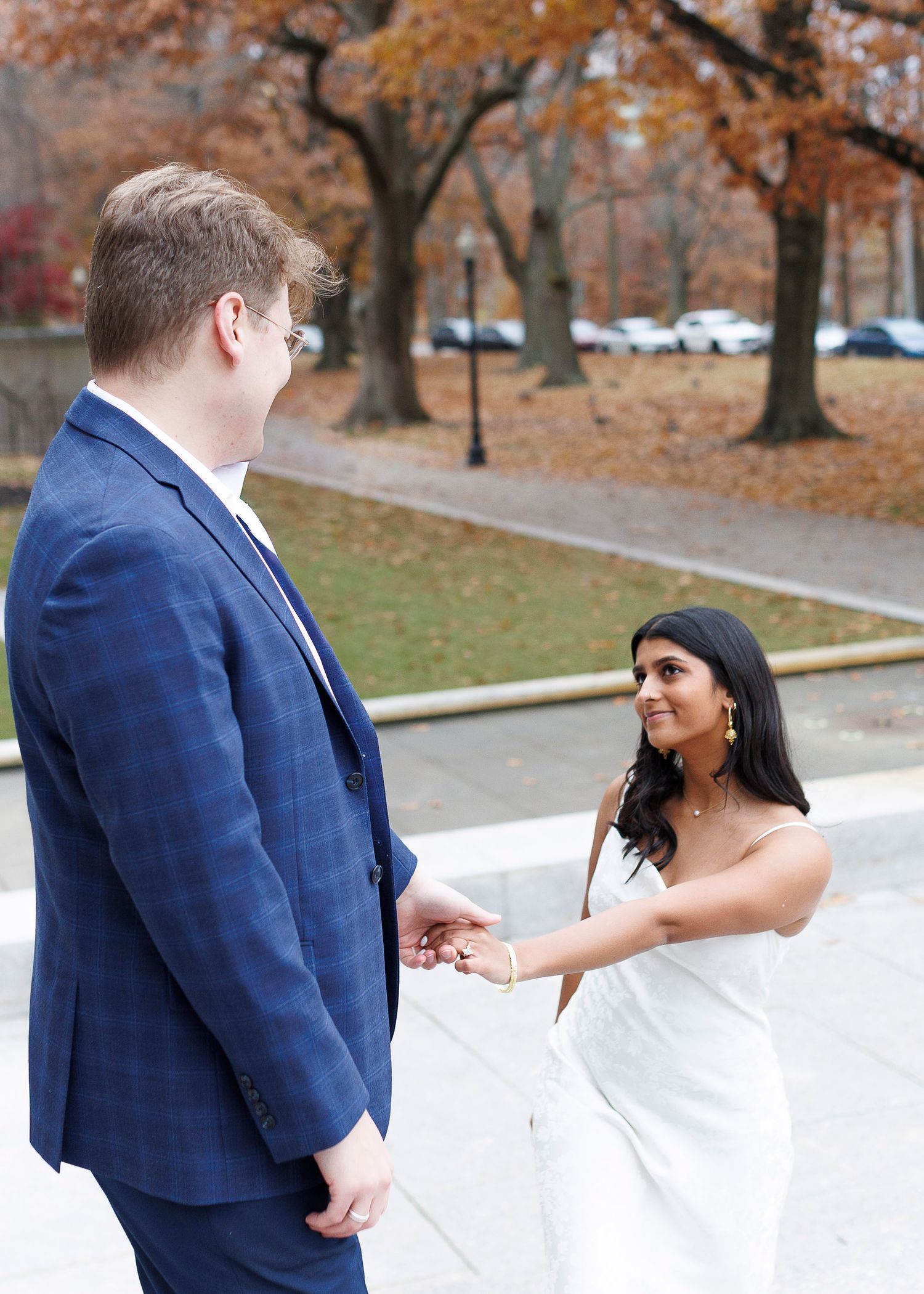 A couple in wedding attire shares a joyful moment outdoors on an autumn day with fallen leaves in the background.