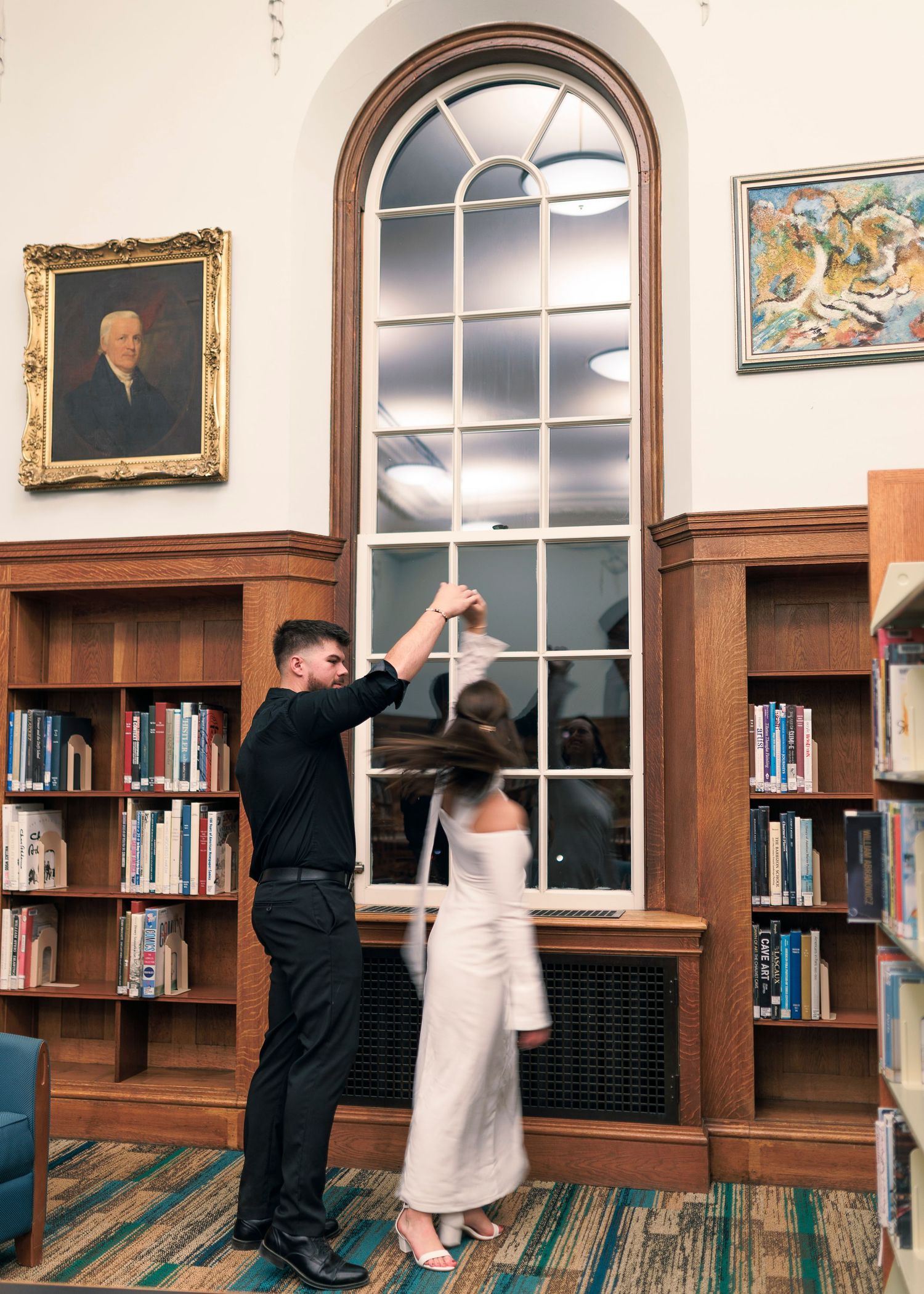 A couple dances together in a library with tall arched windows and wooden bookshelves lining the walls at Brookline Public Library.