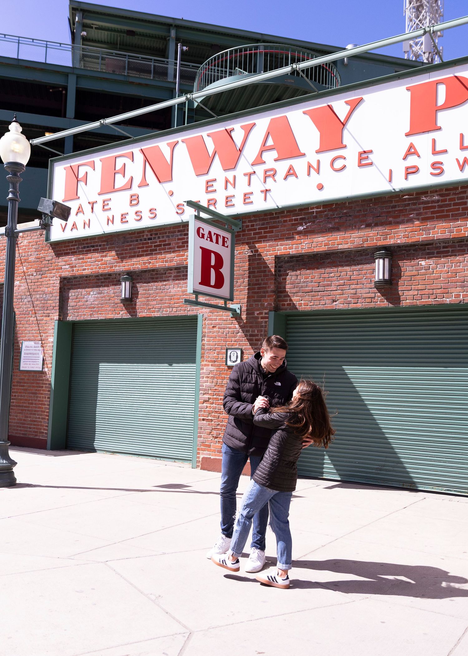 Two people embrace outside Gate B at Fenway Park in Boston on a sunny day.
