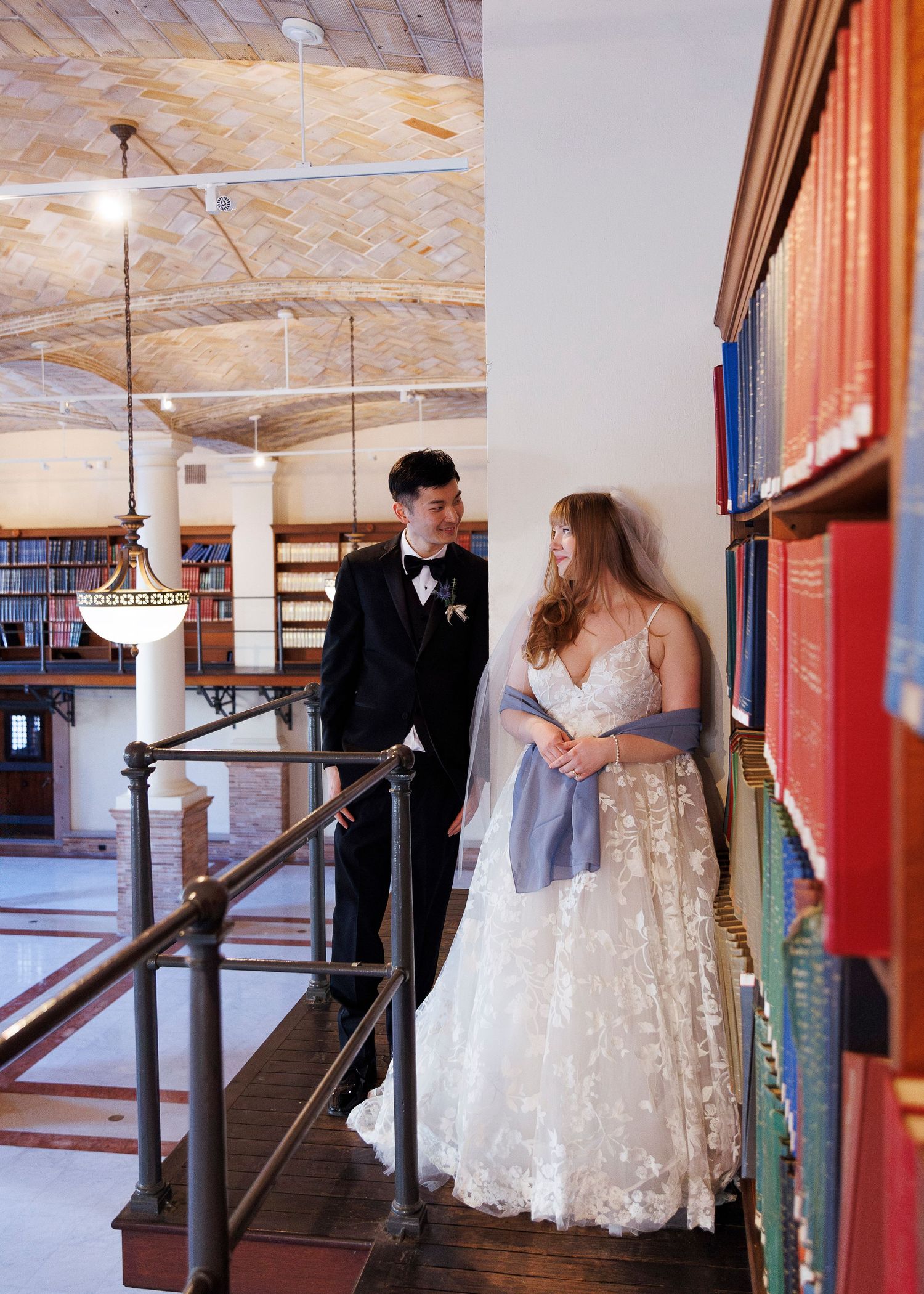 A couple in formal wedding attire poses together on a rustic wooden staircase in an indoor venue.