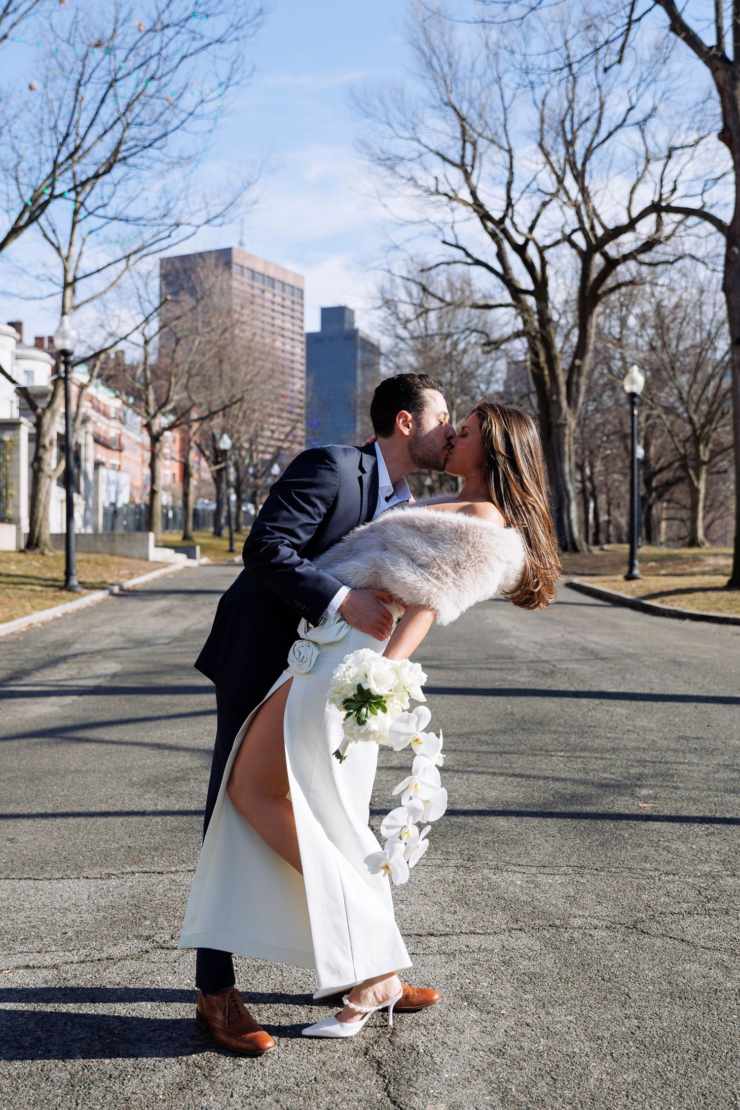 A romantic couple in wedding attire share a kiss on a city path lined with bare trees and brick buildings.