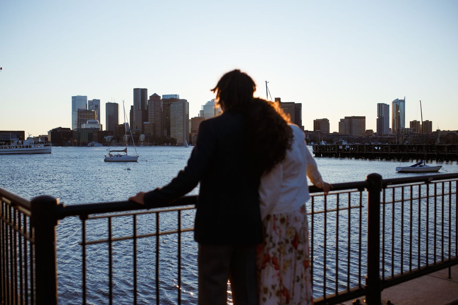 Silhouetted figures stand along a waterfront railing with city skyline and boats visible in the background at sunset.