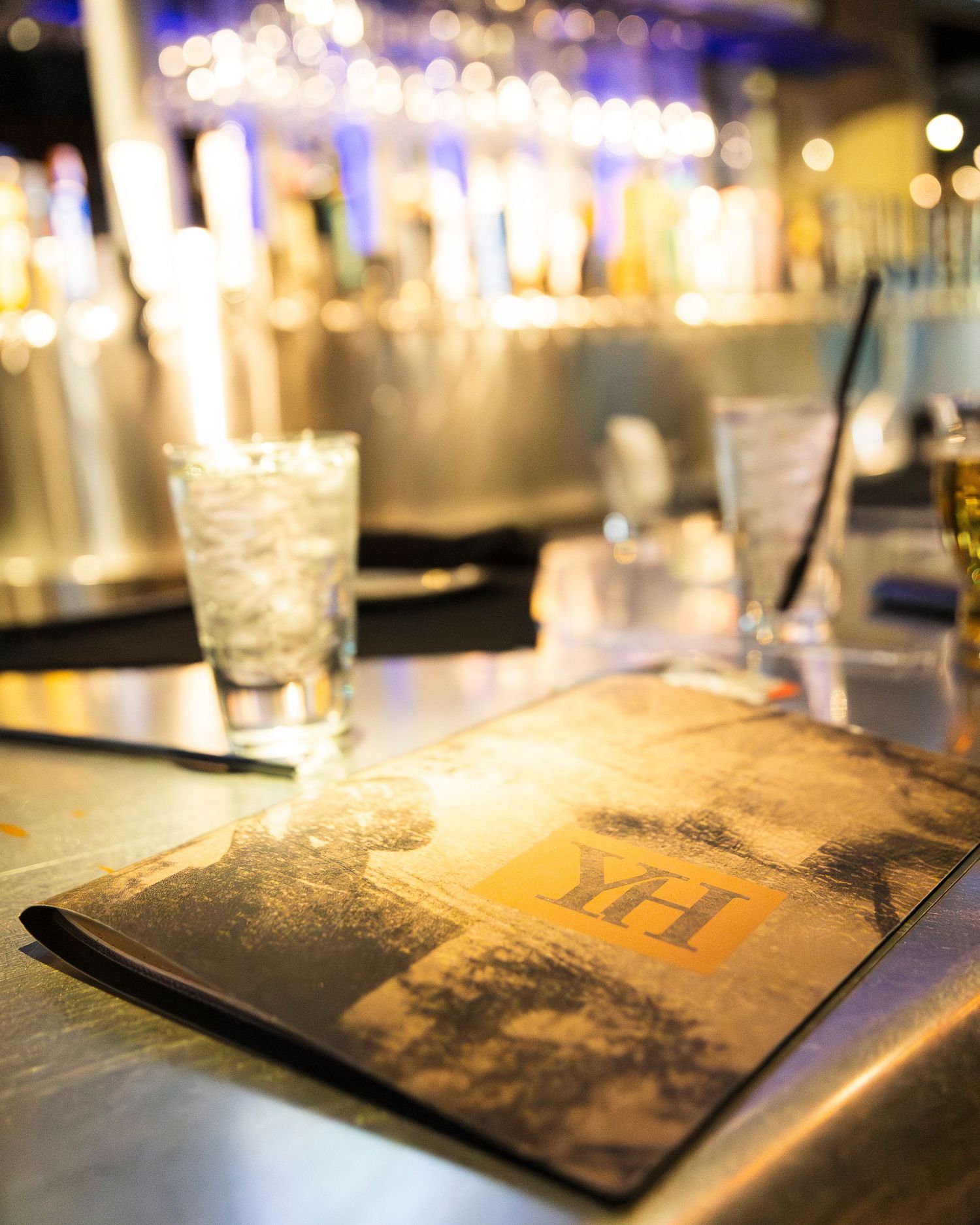 A YH branded menu rests on a bar counter beside glasses of ice water with bokeh bar lights in background.