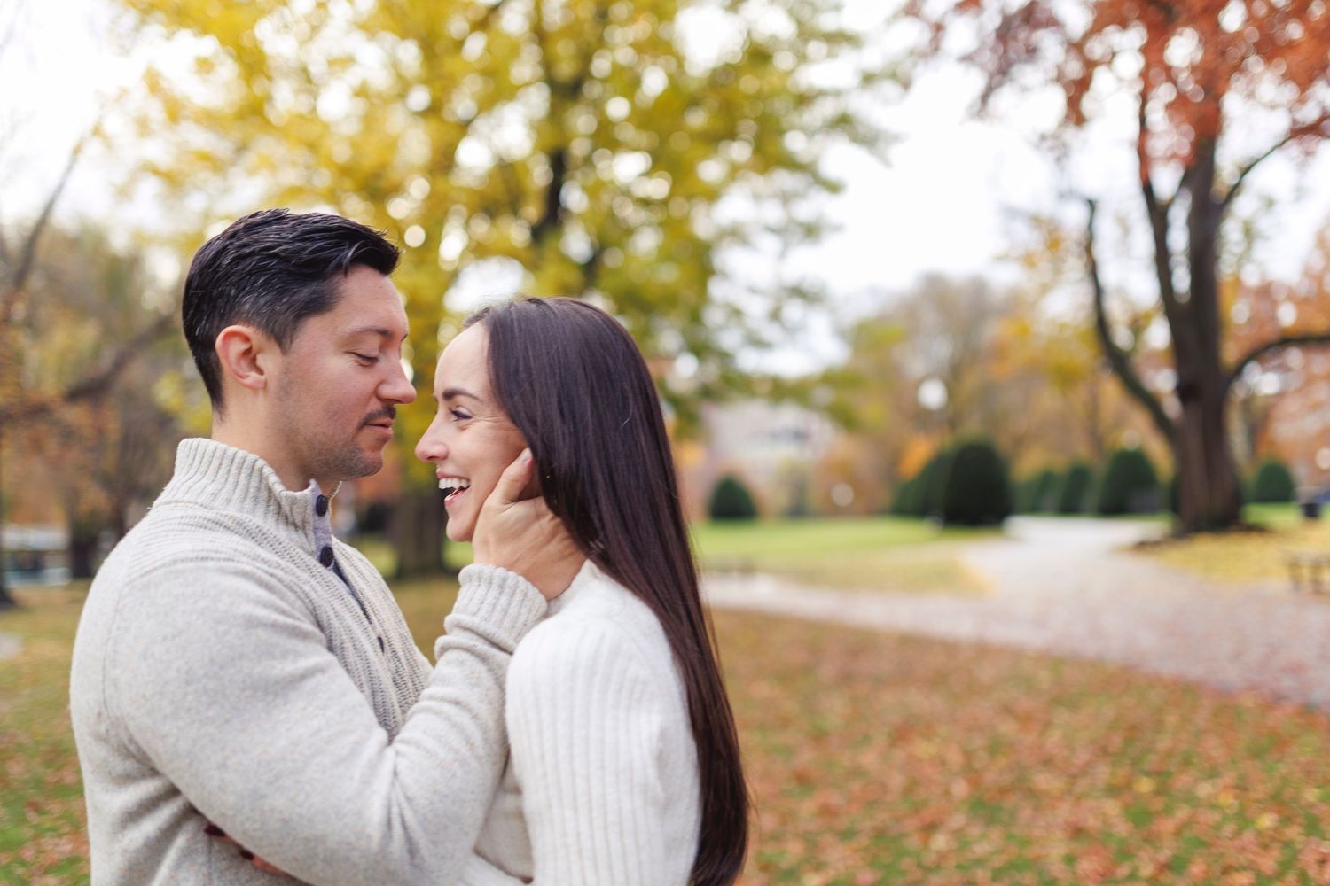 A couple shares an intimate moment outdoors during autumn with fallen leaves and trees in the background.