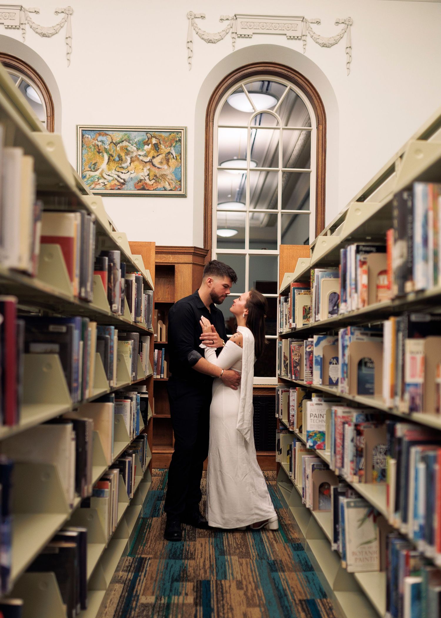A couple shares a romantic moment between library bookshelves, creating an intimate scene in the quiet library setting.