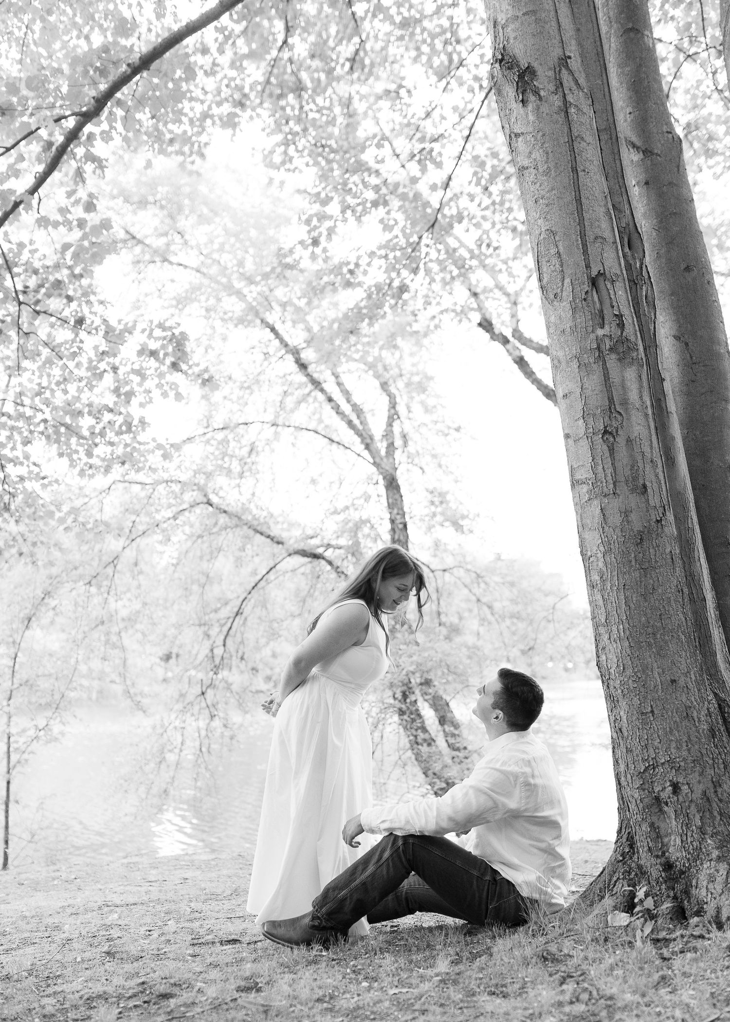 Black and white maternity photo of a pregnant woman in white dress with partner seated under a tree by a lake.
