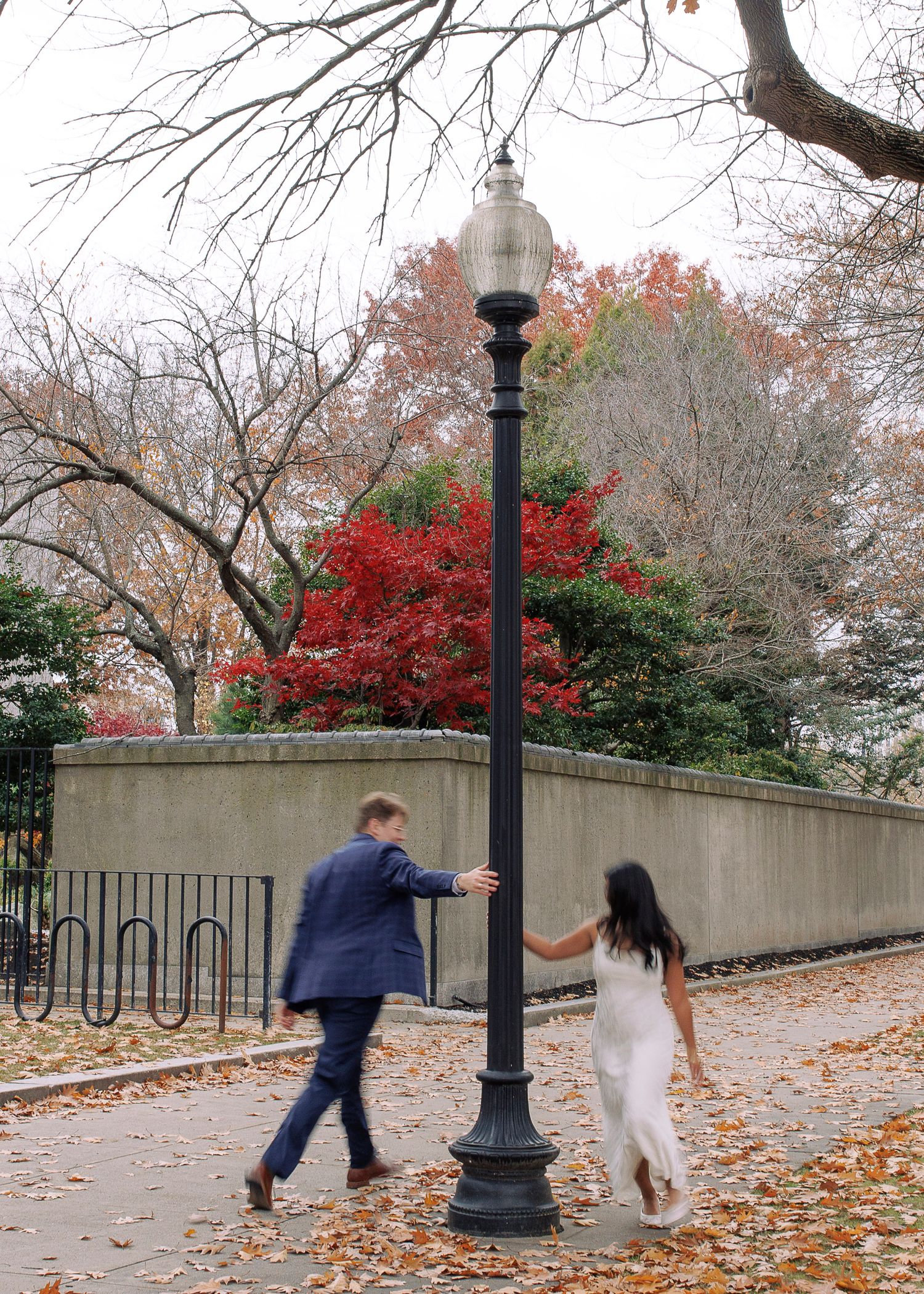 A couple playfully chases each other along a leaf-covered walkway with lamp posts and autumn foliage in the background.