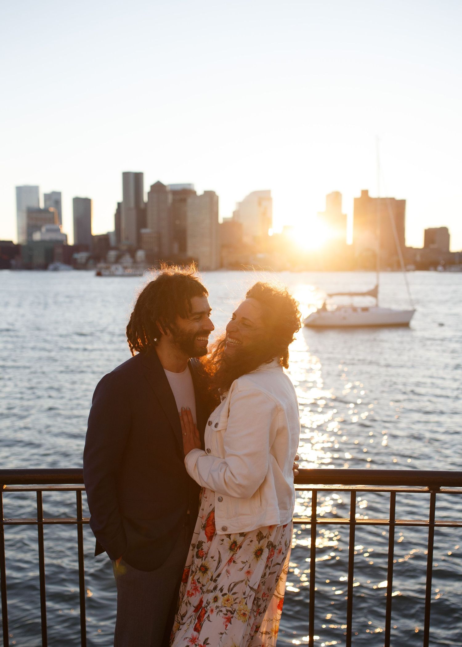 A romantic couple shares a tender moment at sunset by the harbor with boats and city skyline in the background.