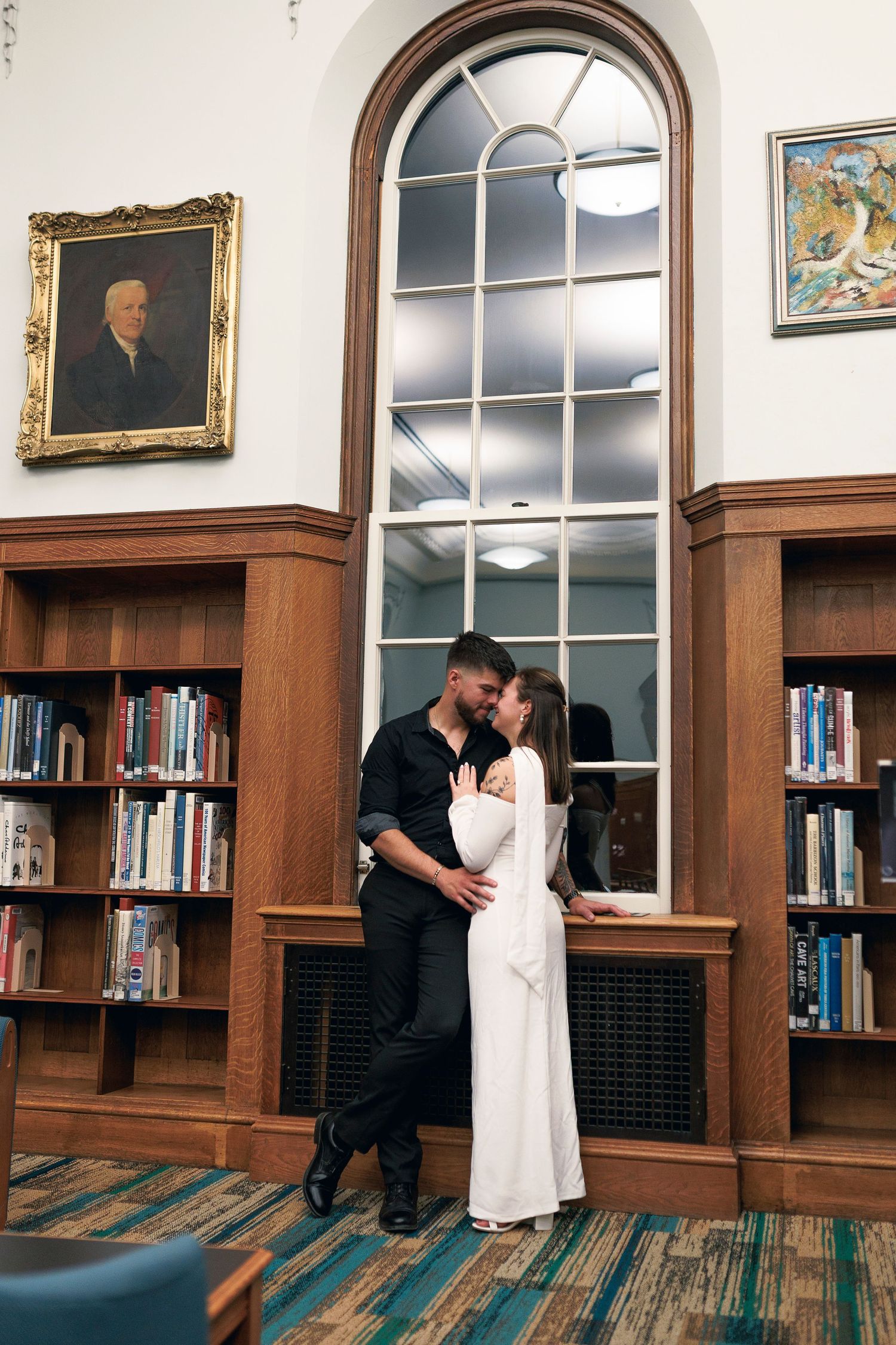 A couple embraces in an elegant library with tall arched windows, wooden bookshelves and vintage decor.