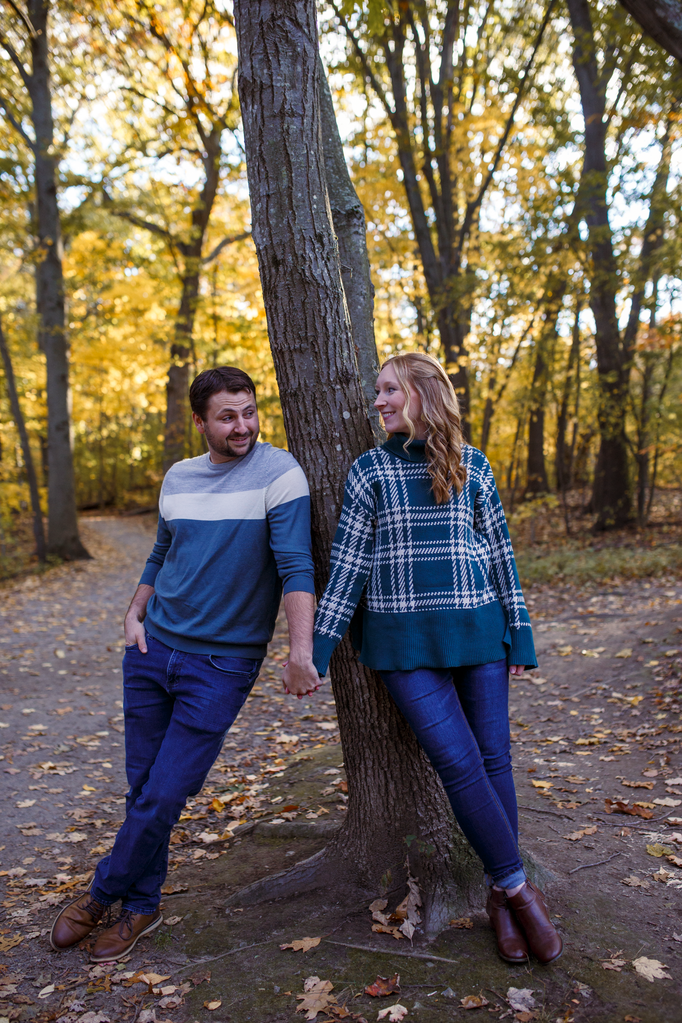 A couple leans against a tree trunk in an autumn forest with golden foliage and fallen leaves on the ground.