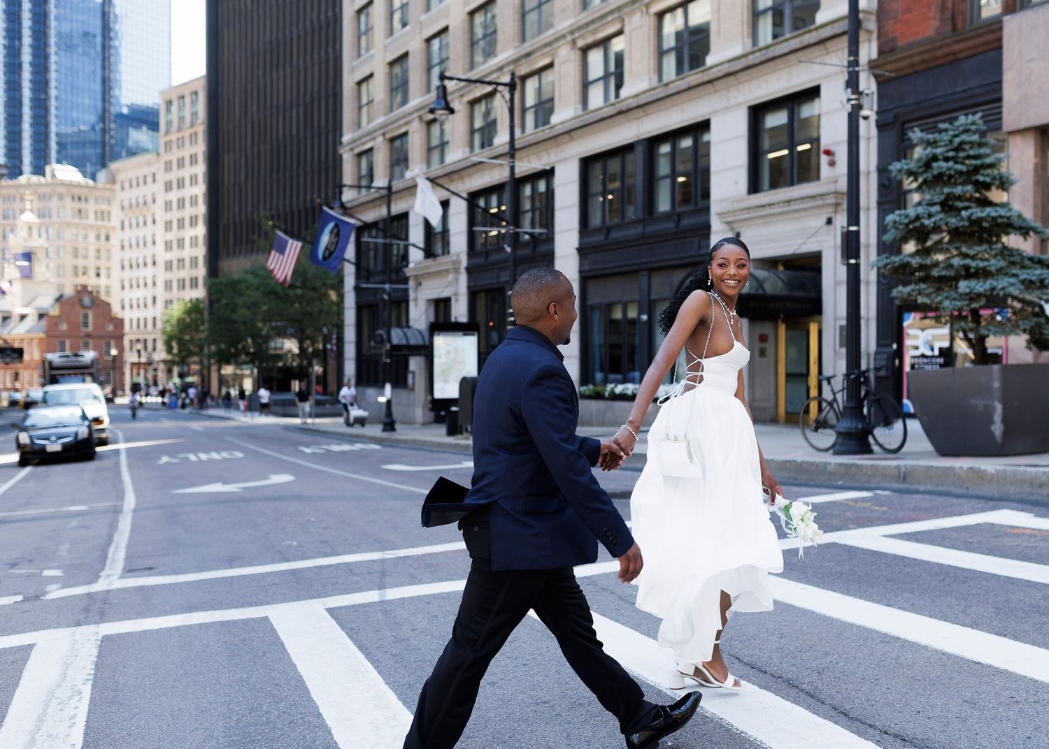 A bride and groom cross a city street in formal wedding attire, with urban buildings and an American flag in the background.