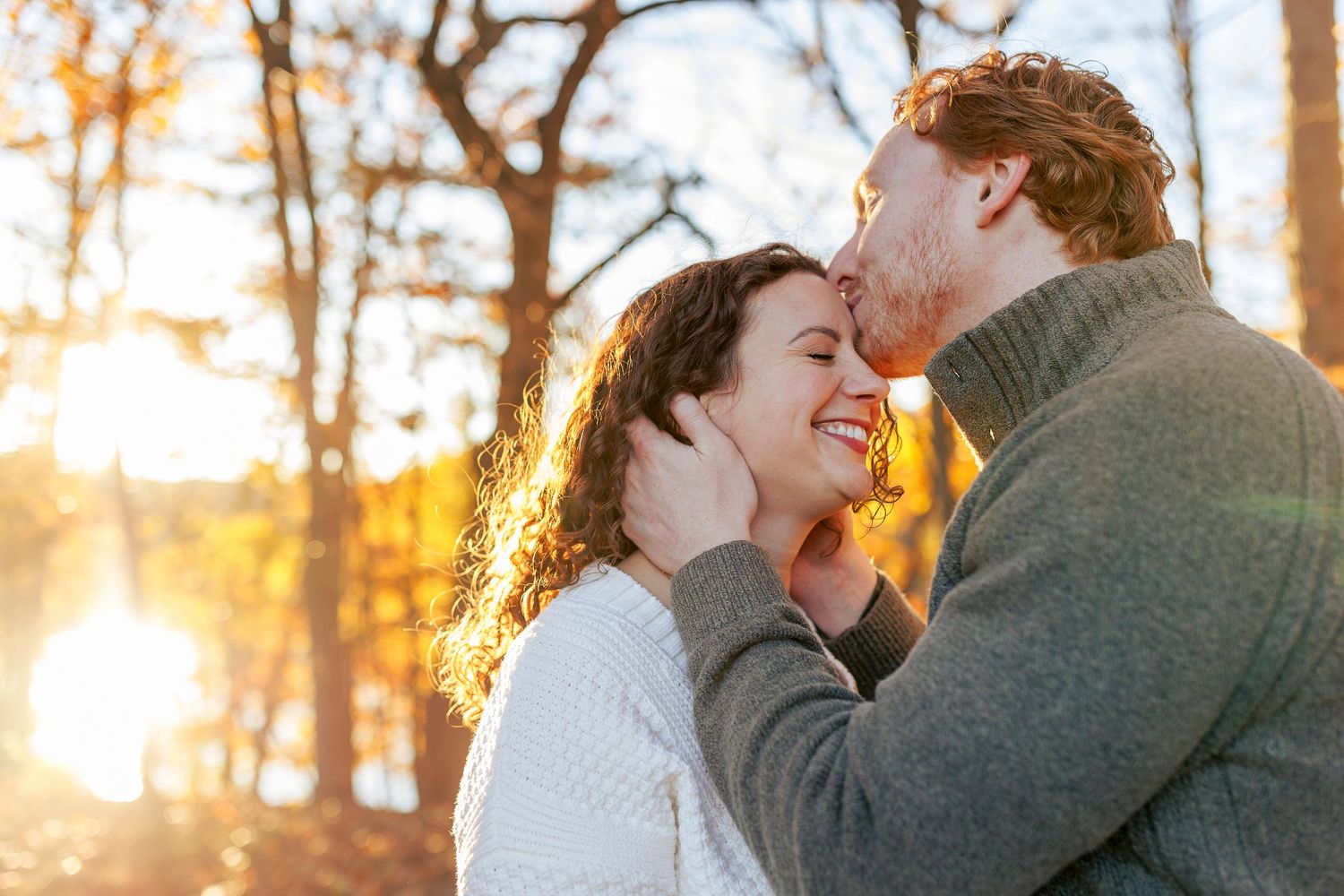 A couple shares a tender moment while laughing together in a sunlit forest during golden hour.