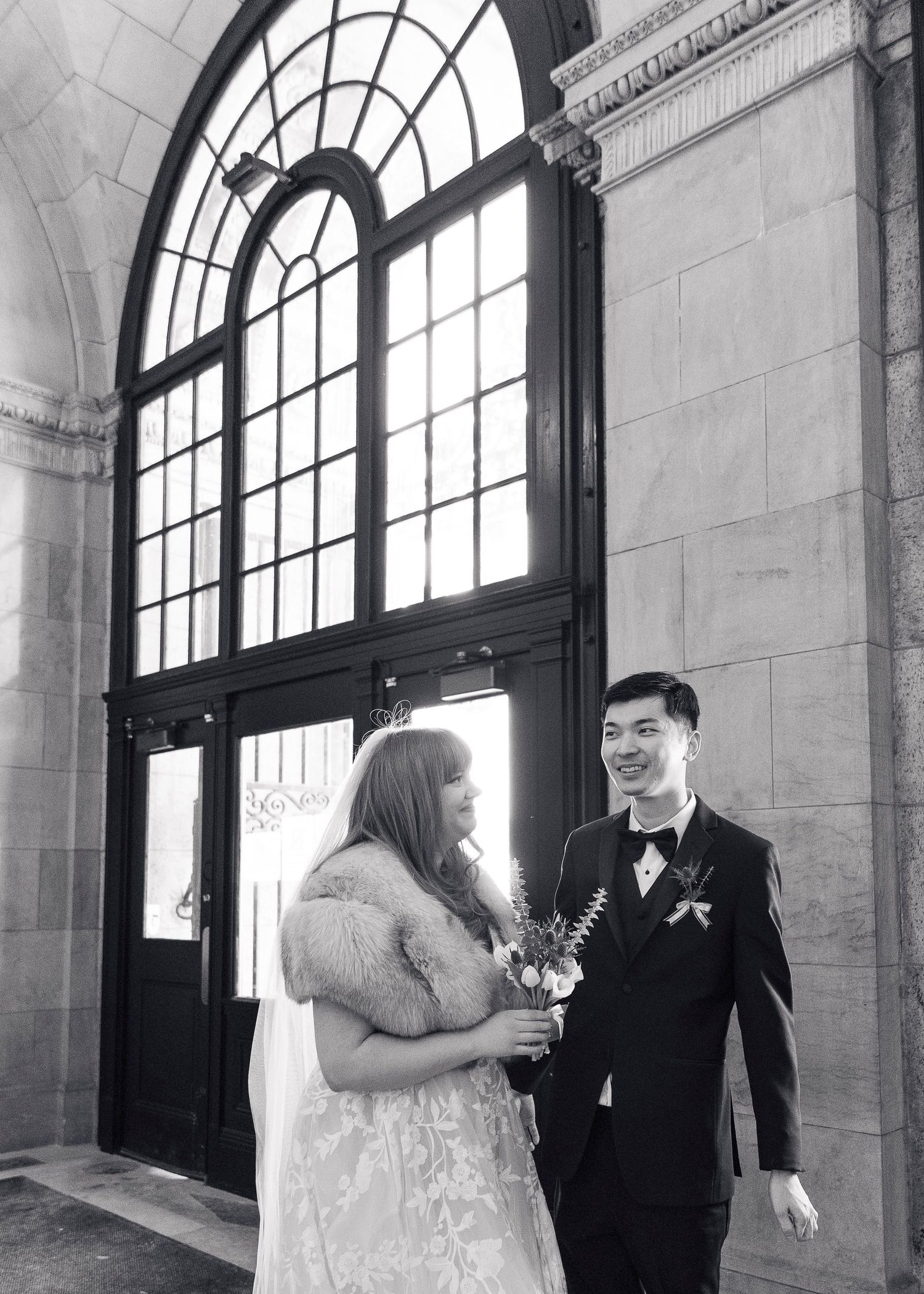 A wedding couple shares a moment under a grand arched window in a historic building.