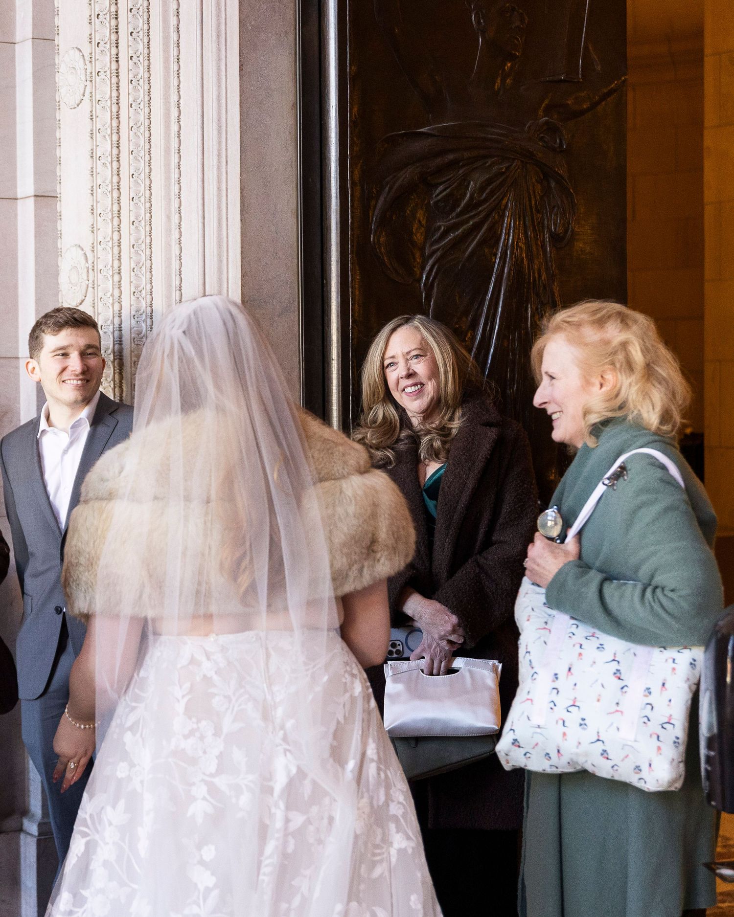 Wedding party members gather in a grand hallway with ornate architectural details and natural lighting.