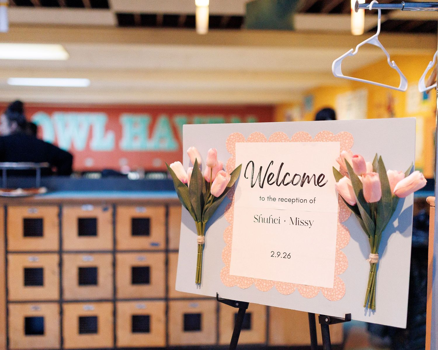 A welcome sign hangs with pink tulips against a blurred retail store background with wooden storage cubbies at Flatbreads in Somerville.