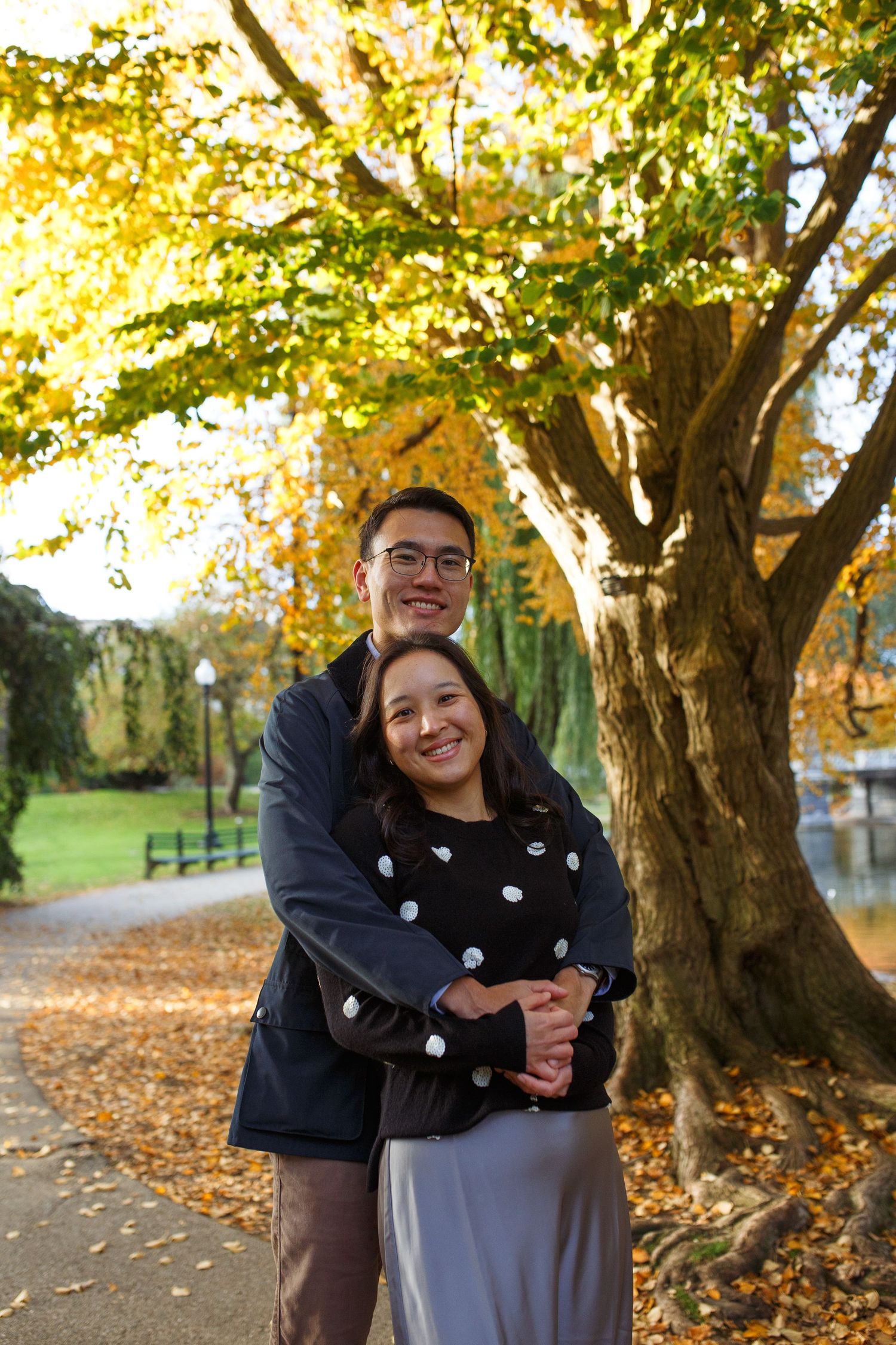 A couple embraces outdoors on an autumn day with golden leaves and trees in the background.