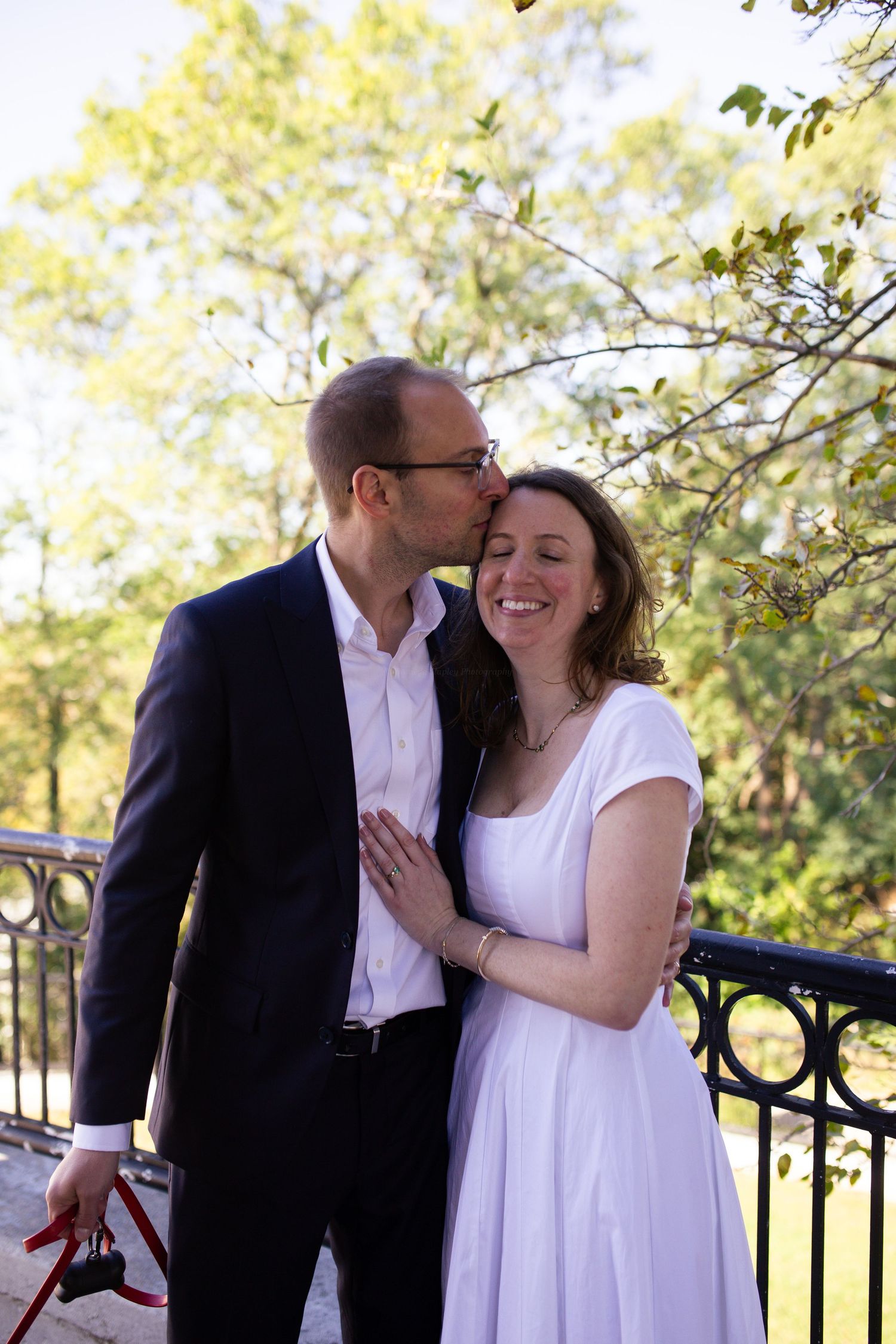A couple in white clothing share tender moments on a balcony surrounded by green foliage on a sunny day.