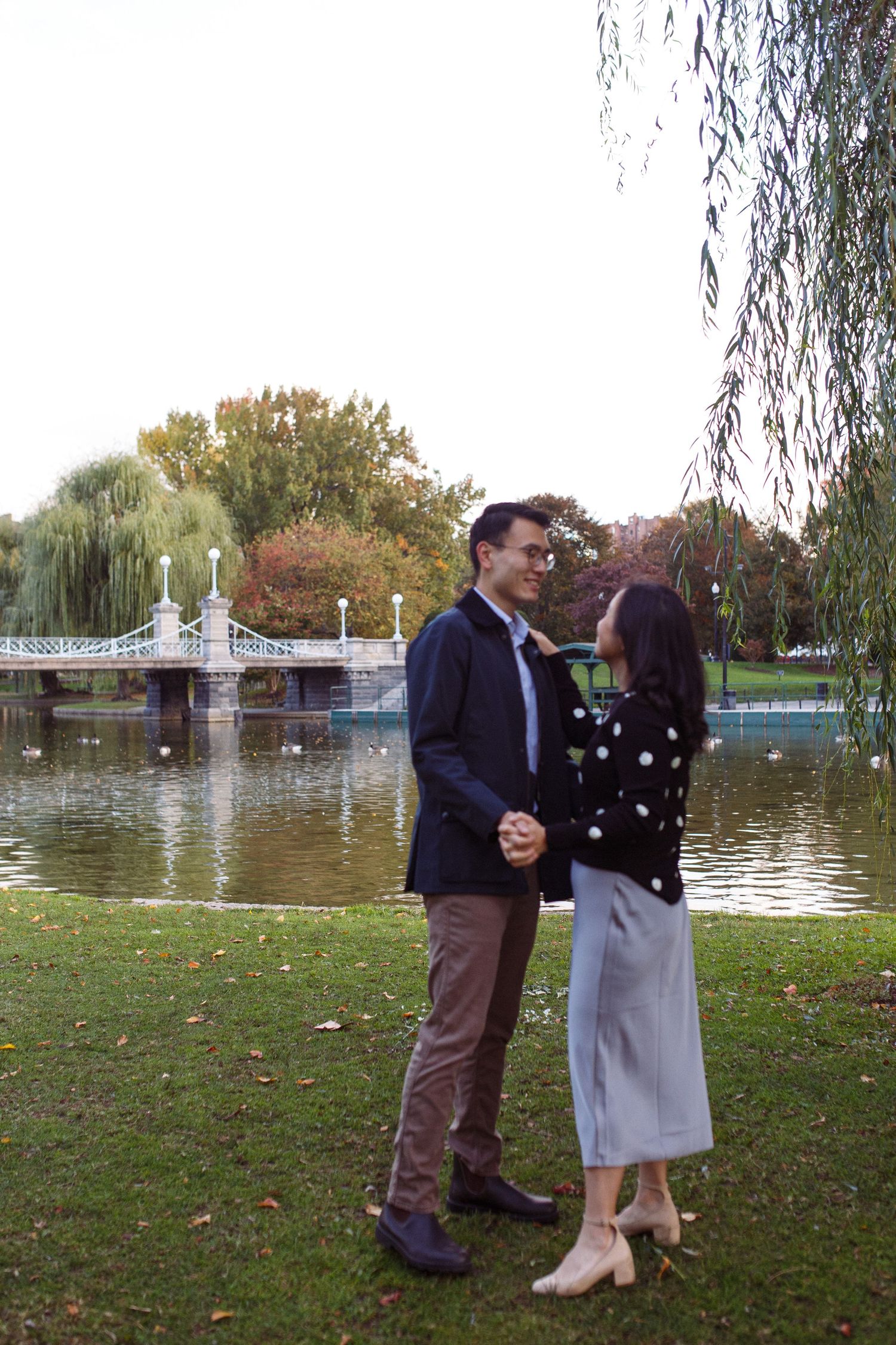 A romantic couple shares a tender moment by a pond with weeping willows and a bridge in a scenic autumn park setting.