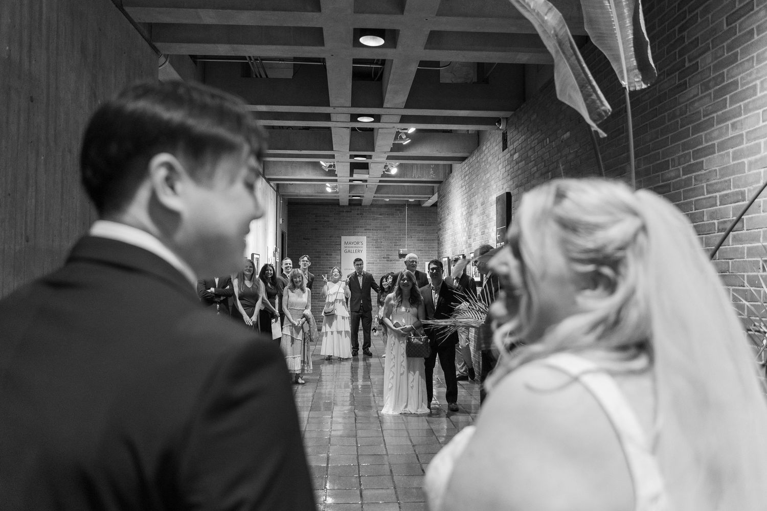 Black and white wedding photo of bride and groom walking toward guests gathered in a brick hallway.